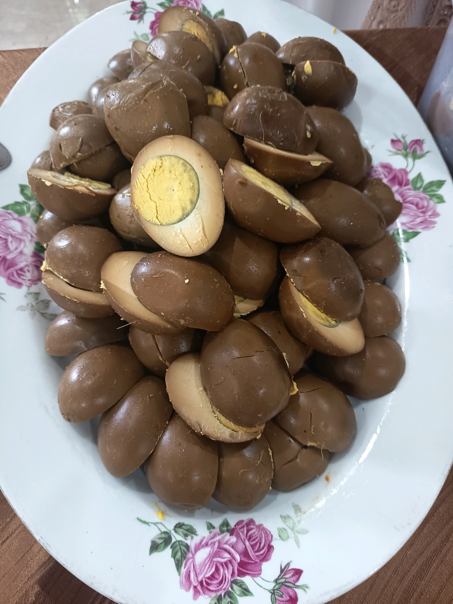 The image shows a white plate with a floral pattern, filled with brown eggs. Some of the eggs are whole, while others are halved, revealing their yellow yolks. The eggs appear to be cooked and seasoned, likely in a soy-based sauce. The plate is placed on a brown, textured surface, possibly a tablecloth. The background is slightly blurred, but suggests an indoor setting, likely a dining table. The image is taken from a top-down perspective. There are no people or specific locations within the city of Bogor, Indonesia, identifiable in the image.
