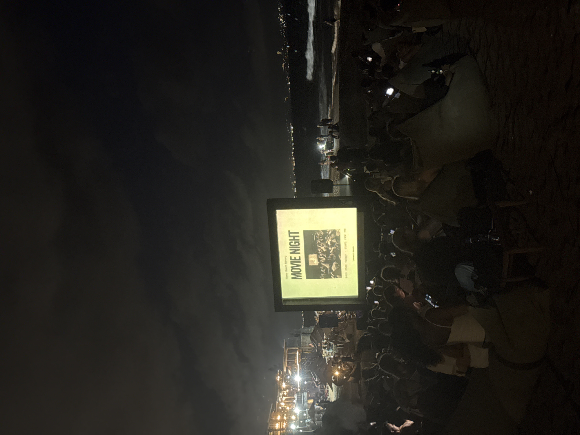 At night in an unidentified city, a large crowd gathers on the beach for an outdoor movie night. People are seated on chairs and beanbags facing a screen that says "Movie Night." Many are holding up cell phones, seemingly recording or taking pictures of the screen. Lights from buildings are visible in the background along the shoreline. The sky is dark with some visible clouds.