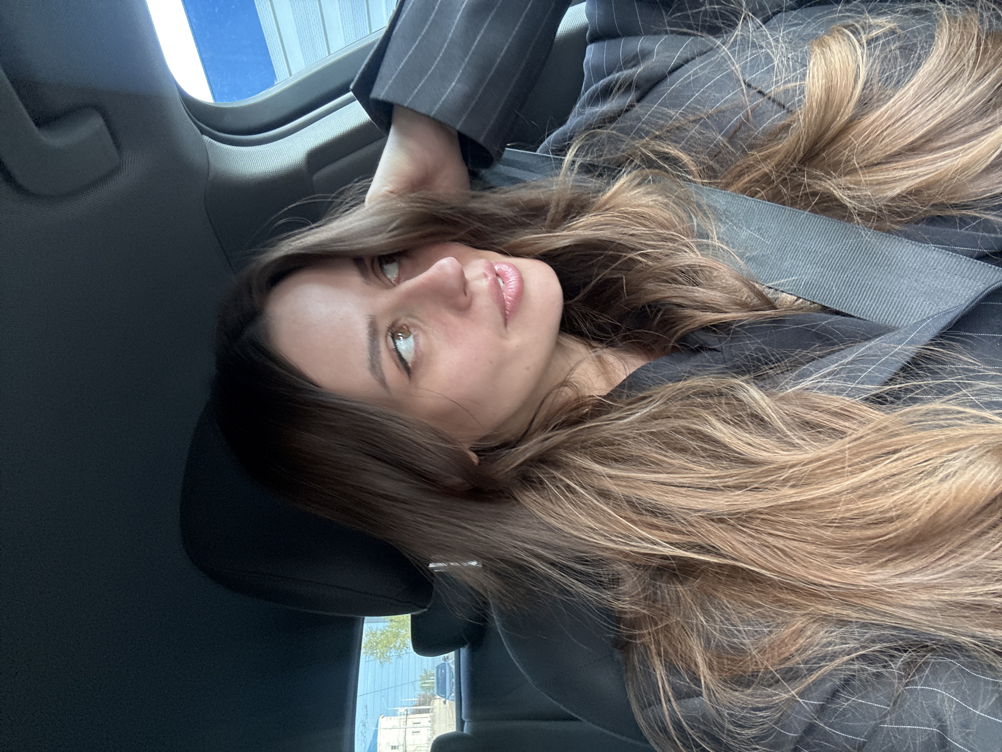 A woman with long brown hair is sitting in a car. She is wearing a dark pinstripe suit jacket and a seatbelt. The background shows a view through the car window, including buildings and a blue sky. The location appears to be an urban area. It is not possible to determine if the image was taken in Krasnoyarsk, Russia from the provided information.