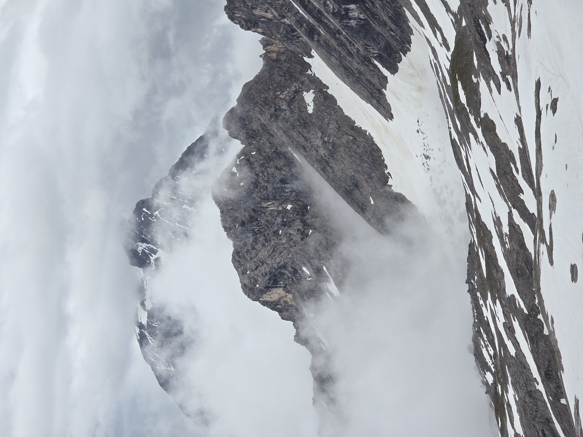 This is a vertical, outdoor, medium shot photograph of the Zugspitze, Germany's highest peak, located near Garmisch-Partenkirchen. The scene is characterized by rugged, rocky mountain slopes partially covered in snow. Wisps of fog and clouds drift across the landscape, obscuring parts of the mountains and creating a dramatic, atmospheric effect. The foreground shows a steep, rocky incline with patches of snow, transitioning to more substantial snowfields in the middle ground. Several jagged, dark grey mountain peaks rise in the background, their summits partially shrouded in clouds. The overall color palette is muted, dominated by greys, whites, and dark rock tones, conveying a sense of cold and isolation. There are no people or visible structures in the frame.