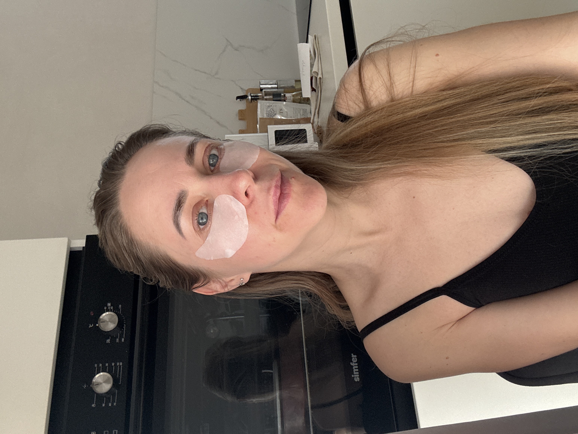 A portrait-oriented image shows a young woman engaging in a skincare routine in what appears to be a modern kitchen. The woman, with light brown or blonde long hair, blue eyes, and fair skin, is looking directly at the camera with a neutral expression. She has pink-toned hydrogel eye patches applied under both eyes. She is wearing a black tank top or camisole with thin straps.
The setting is indoors, likely a kitchen or utility area, suggested by the presence of appliances. To her left, partially visible, is a built-in oven or similar appliance with control knobs and a reflective glass door. On the glass panel, the brand name "Simfer" is visible. Above the oven, a white tiled wall with subtle grey veining extends into the background. Behind her, on a counter or shelf, various bottles, jars, and other personal care or cosmetic items are arranged, though mostly out of focus.
The overall impression is one of a casual, self-care moment at home. The image appears to be a selfie or a candid shot. Based on the provided context, the location is Chaikovsky, Russia.