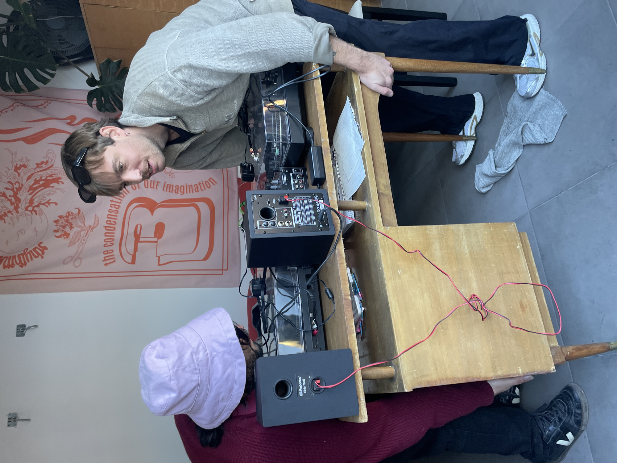 Two individuals are gathered around a light-colored wooden two-tiered stand that appears to hold electronic equipment, including a turntable, audio mixer, and speakers. The person on the left is wearing a light-colored jacket and has sunglasses pushed up onto their head. They are looking down at the equipment. The person on the right is wearing a maroon sweater and a light pink bucket hat. The equipment is connected by red cables. The scene is indoors, with a concrete floor and a decorative banner hanging on the wall with text and images. A plant is visible in the background. Outside of that no information is available from this image to imply that this is within the city of Buenos Aires, Argentina.