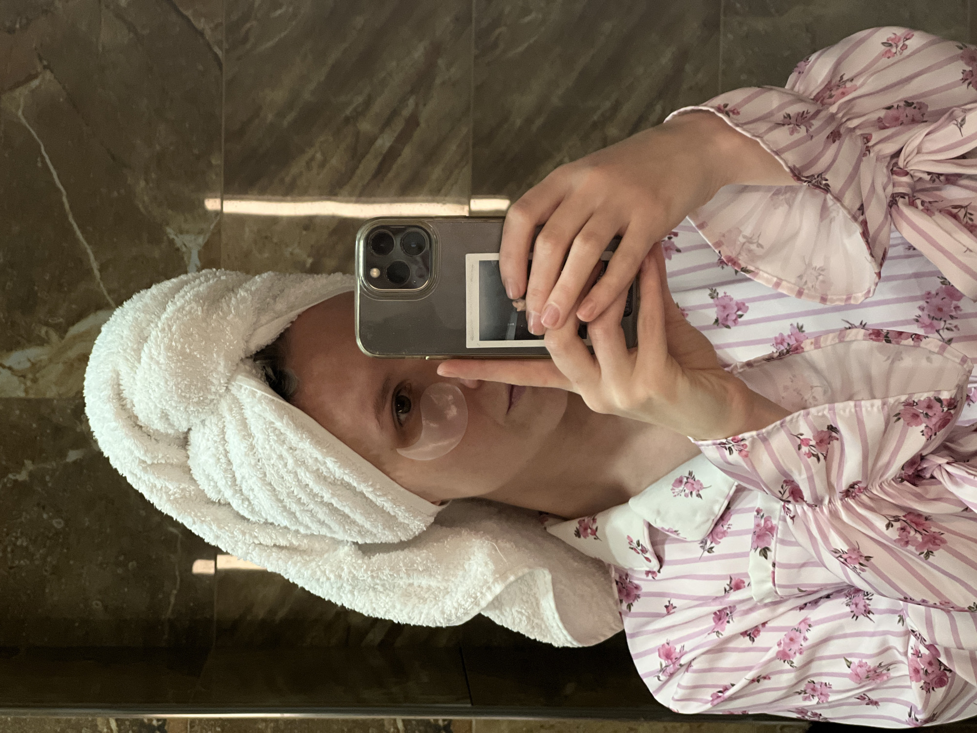 A person is taking a selfie in front of a mirror. They are wearing a pink and white striped shirt with floral patterns and a white towel wrapped around their head. A gel eye mask is under their left eye. They are holding a phone with multiple camera lenses in their hands. A photograph is attached to the back of the phone. The background appears to be a brown marble or tiled wall. There is no evidence to suggest that this image was taken in Khimki, Russia.