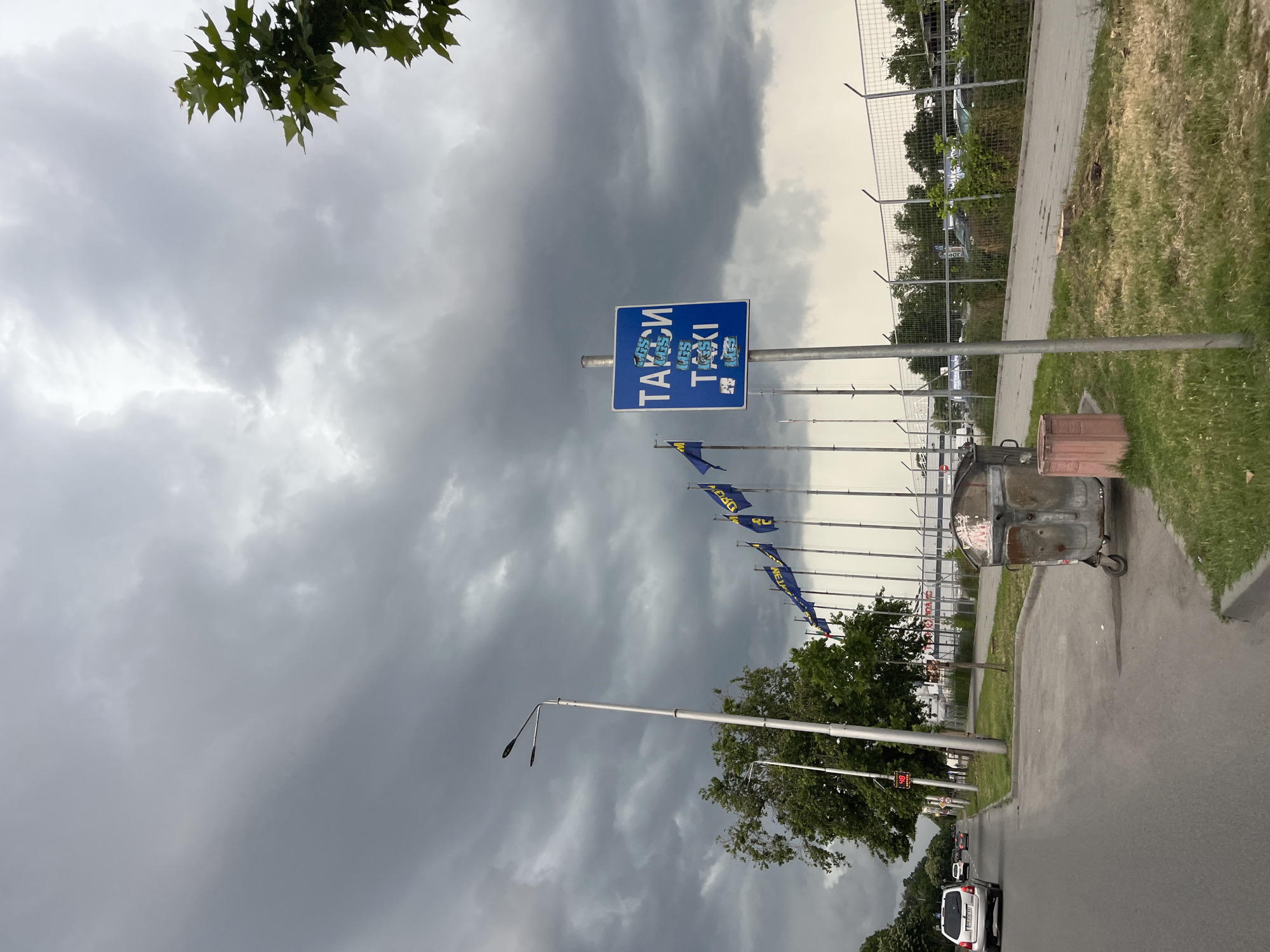 The image depicts an outdoor street scene in Plovdiv, Bulgaria, characterized by an overcast and heavy sky dominated by dark grey clouds. The foreground features an asphalt road on the left, where a silver SUV with the Bulgarian license plate "P08 47 AA" is visible, along with other vehicles receding into the distance. A red digital speed display showing "48 KM/H" is positioned on a pole further down the road.
To the right of the road, a paved sidewalk borders a strip of grass. A weathered metal dumpster on wheels is situated on the grass, adjacent to a pinkish concrete bin or planter. A blue rectangular sign on a pole, reading "TAKSI" in Cyrillic and "TAXI" in Latin script, is prominently displayed, partially obscured by multiple small stickers.
Behind the grass and signage, a series of tall poles bear blue and yellow flags, some exhibiting yellow lettering consistent with the IKEA logo. These flags stand in front of a substantial facility enclosed by a tall metal fence topped with barbed wire, suggesting a commercial or industrial area. Trees with green foliage are scattered throughout the scene, including a prominent branch in the upper right corner and a line of trees along the left side of the road. Multiple streetlights with double luminaires are spaced along the roadside. The overall environment suggests a commercial district or the vicinity of a large retail outlet, specifically near the IKEA store in Plovdiv.