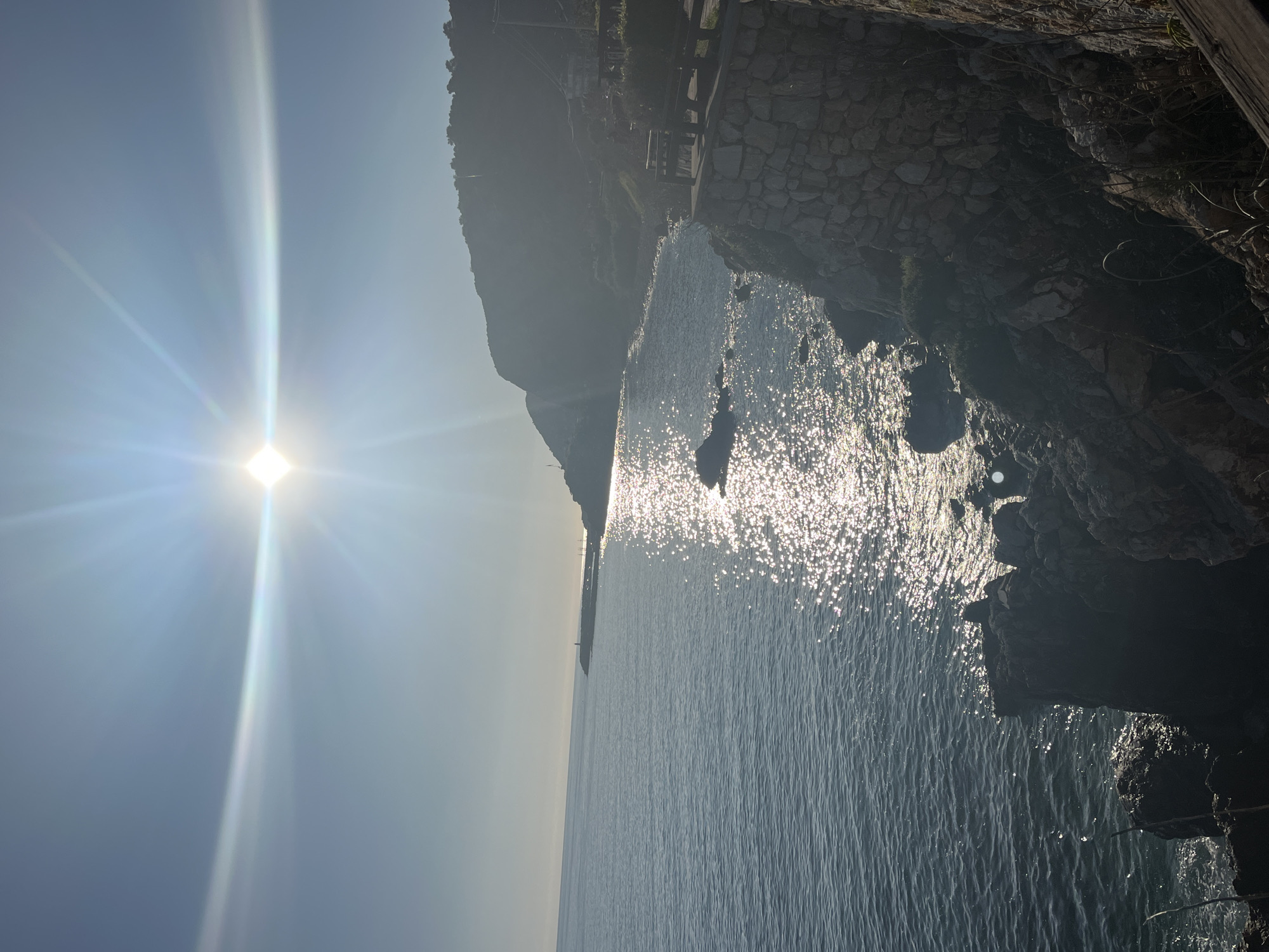 The image displays a bright coastal landscape under intense sunlight. The scene is primarily composed of a large body of water, likely the sea, occupying the lower and middle-right portions of the frame. On the far left, a brilliant, radiant sun, appearing almost diamond-shaped, is prominent, casting a strong lens flare and creating a shimmering, sparkling path of light across the water's surface. The water exhibits numerous small ripples, reflecting the sunlight in countless glinting points.
To the right, a steep, rugged, and dark rocky cliff face rises sharply from the sea. This cliff features sparse vegetation, including moss and small plants. Along the upper section of the cliff, man-made structures are visible, including what appears to be a stone wall or pathway, and potentially wooden railings or benches, suggesting a developed coastal trail or viewpoint. Further in the distance, a long, dark silhouette of a landmass or mountain range stretches along the horizon, with a faint, small structure, possibly a lighthouse, discernible on it.
The sky, particularly around the sun on the left, is clear and bright blue, transitioning to a lighter, hazy shade towards the horizon. The high, intense sun indicates it is mid-day or early afternoon. The weather is clear and sunny. There are no people visible, nor are any specific activities taking place; the focus is on the natural grandeur of the coastline. The overall impression is one of serene natural beauty and warmth, characteristic of a scenic coastal location such as Alanya, Turkey. No visible text is present in the image.