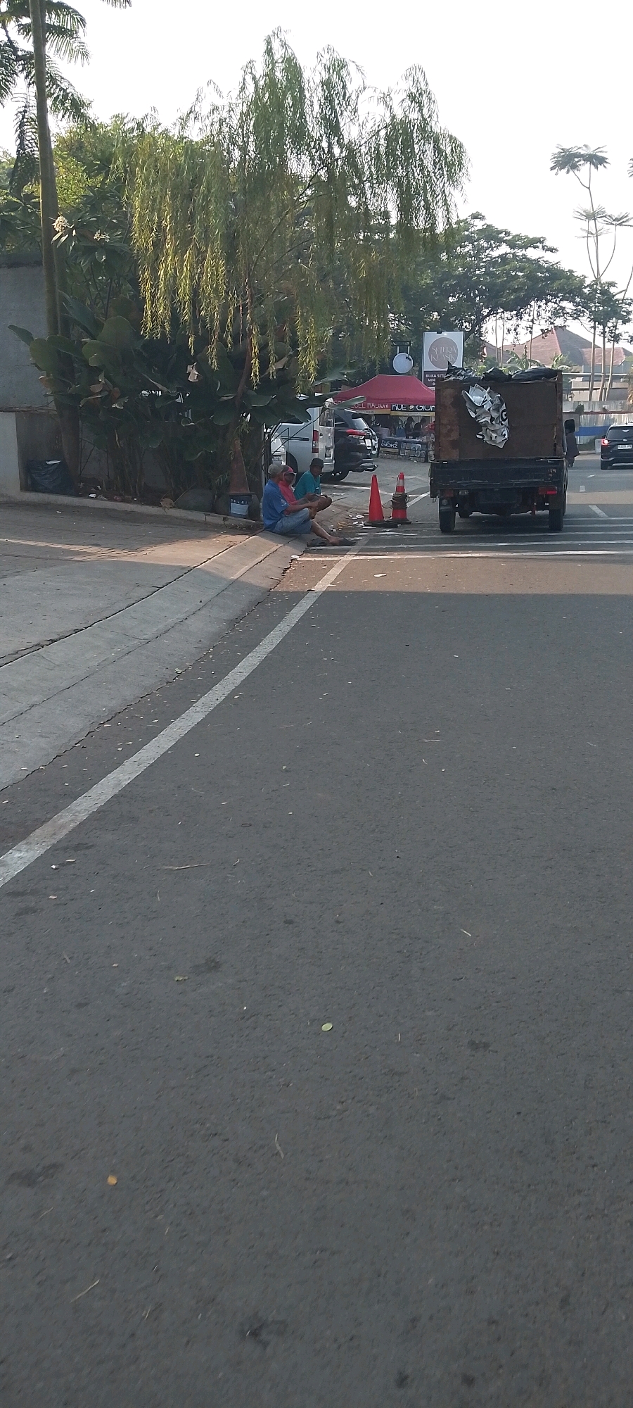 This image depicts a street scene in Gunung Putri, Indonesia. In the foreground, a gray asphalt road is visible with a white line marking the lane division. On the right side of the road, a pedestrian crossing is indicated by white stripes. To the left of the road, a concrete curb separates it from a paved walkway.
Seated on the curb are two individuals. One person, wearing a blue shirt and dark pants, is seated with their legs extended. Beside them, another person, dressed in a blue shirt and lighter-colored pants, is also seated, holding an object in their hands.
Behind the seated individuals, there is a green weeping willow tree with long, drooping branches, and other lush green foliage. Further back, a dark-colored truck with a wooden bed is parked. The truck bed contains debris. In the background, there are several buildings, including one with a red canopy tent and another with a white circular sign. A dark-colored SUV is parked near the buildings. Various traffic cones are placed on the ground near the truck. The sky is bright, suggesting daytime.