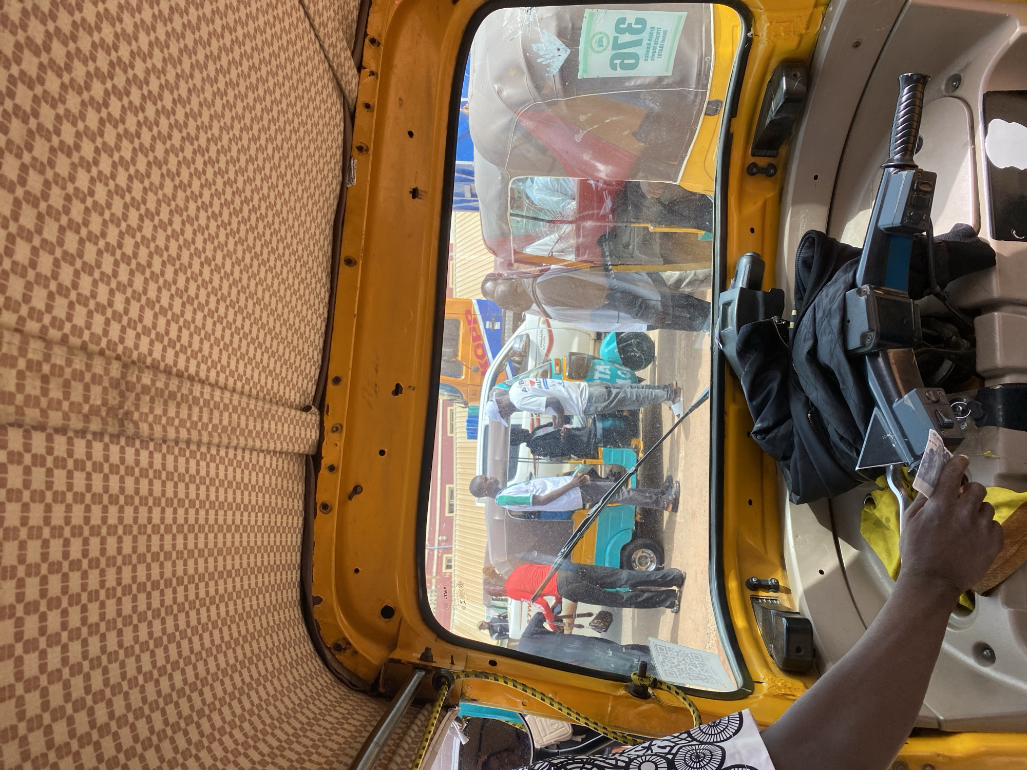 This media file captures an interior view of a yellow auto-rickshaw, also known as a "Keke," likely in Jos, Nigeria. The camera is positioned to give a first-person perspective, looking out through the windshield and side windows.
The dominant subject is the interior of the Keke, with its patterned fabric lining the walls. Outside, visible through the windshield, are other vehicles and people. Specifically, there are several passengers, appearing to be men, seated in what seems to be another Keke or similar vehicle. They are wearing casual clothing, including t-shirts and trousers. One passenger wears a red shirt, another a white shirt with green trim, and another a white shirt with a green collar. Their postures suggest they are traveling.
In the foreground, inside the Keke, a hand is visible holding and seemingly handing over some Nigerian currency. A part of the driver's interface is also visible, including a gear shift or control lever and what appears to be a handle for operating the vehicle. A black jacket is draped over a seat.
The scene appears to be taking place during daylight hours, given the brightness of the image and the reflections on the windshield. The weather seems to be clear, allowing for good visibility.
There is visible text on a sticker on the windshield, which appears to be in a language that is not English, possibly a local language or a stylized logo. Another sticker with a QR code is present on the side of the Keke.
The overall impression is a snapshot of daily transportation and commerce in an urban environment in Nigeria. The activity depicted is likely the completion of a journey or a transaction between passengers and the driver. The setting is clearly an outdoor, public space.