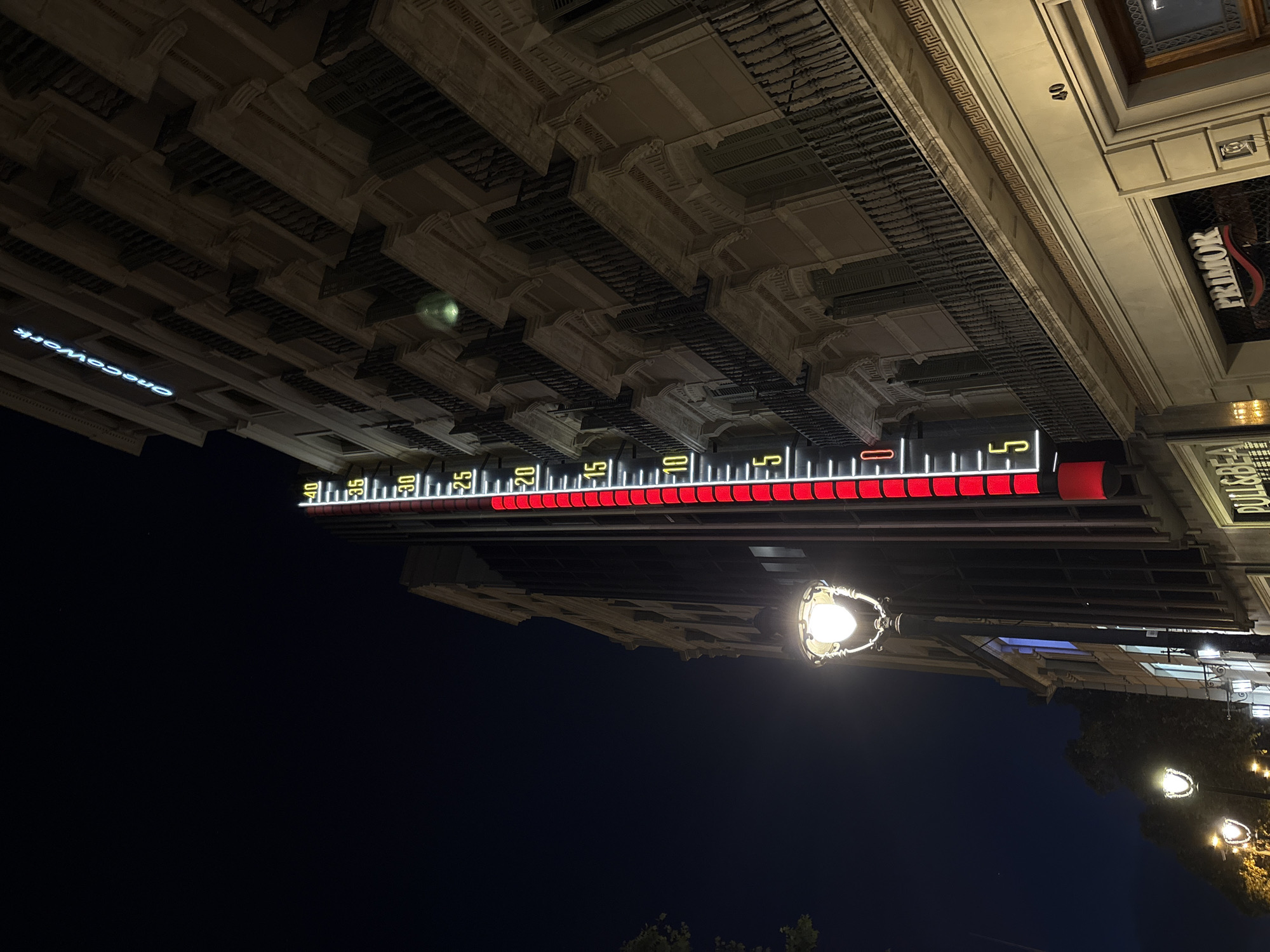 At night, a view of a large, lit thermometer on a building in Barcelona, Spain. The thermometer displays a temperature, visible with a red-illuminated display. Overhead architectural details of the building can be seen along the upper portion of the image. Streetlights are visible below, and the dark night sky fills the bottom half of the frame. Signs for storefronts like “PRIMOR” and “onecowork” are seen on the building.