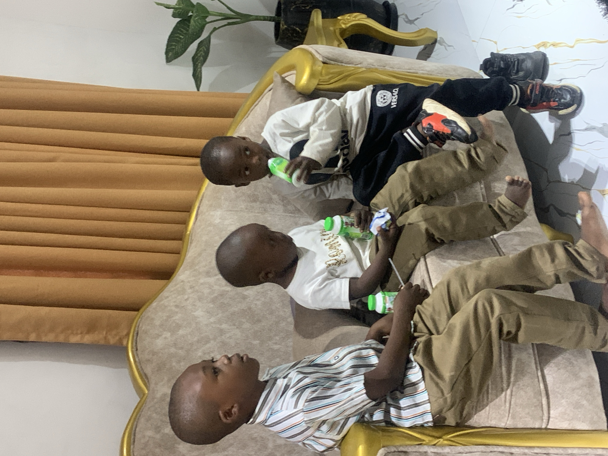 Three young boys are lying on a gold-trimmed sofa. The top boy wears black and white athletic wear with black pants, and has orange sneakers. The middle boy wears a white t-shirt and tan pants. The bottom boy wears a striped collared shirt and tan pants. All three boys are holding green and white bottles. The sofa is placed near a window with a tan curtain, and a plant sits on a stand behind the sofa. The location is Jos, Nigeria.