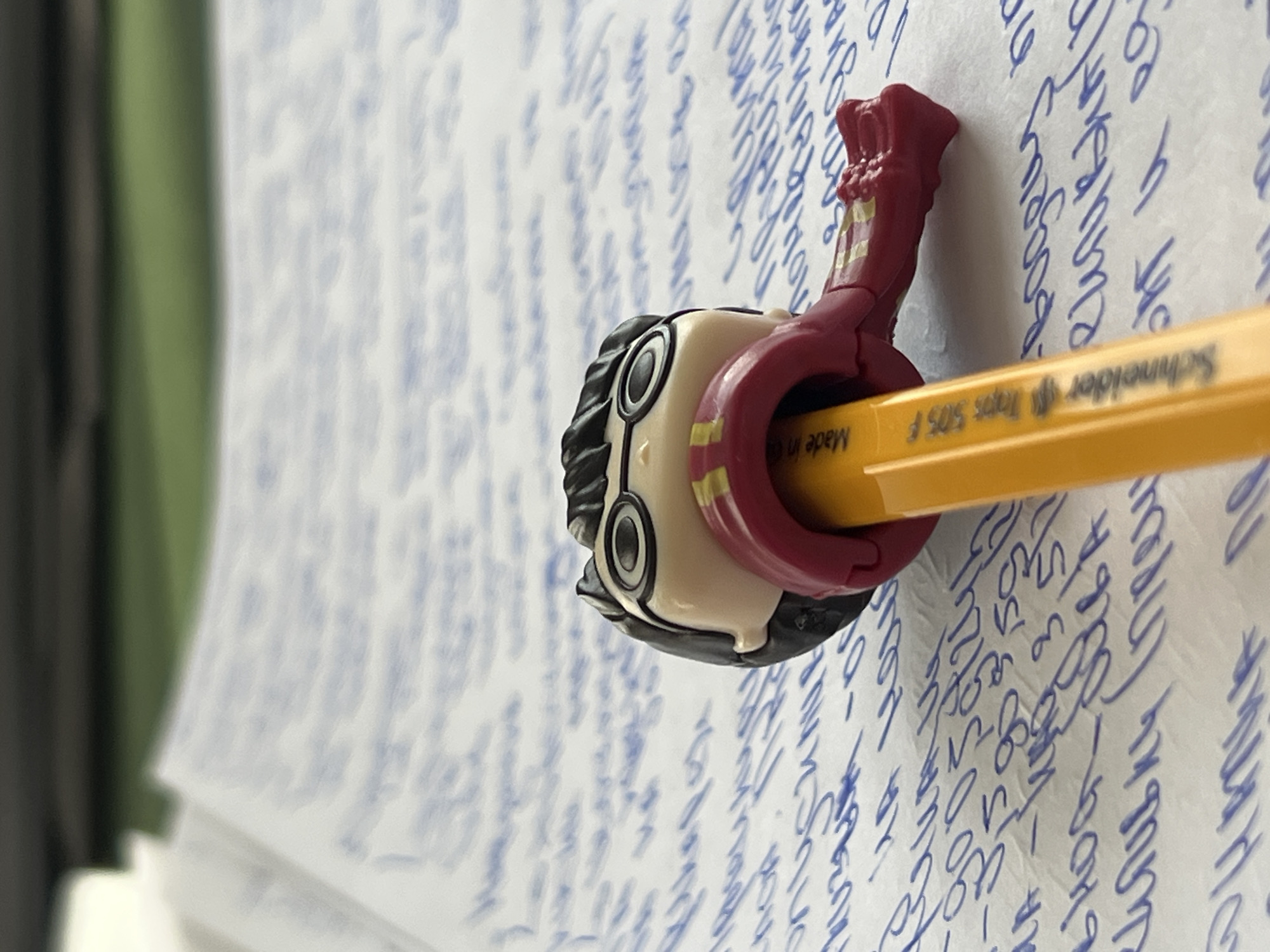 The image displays a close-up, eye-level view of a yellow pencil with a Harry Potter-themed figurine attached to it. The figurine, a character from the Harry Potter series, is positioned as if it is biting or holding the pencil. The pencil itself has visible text printed on it, reading "Made in G Schneider ggg 505 F". The pencil and figurine are resting on a piece of paper covered in handwritten text in blue ink. The background is blurred but suggests an indoor setting with a dark surface at the top and a green area below. The lighting appears to be from above, casting a slight shadow. The image does not provide any specific geographical context or landmarks that would allow for the identification of a location in Burgas, Bulgaria, or any other city. The scene is static and focuses solely on the pencil, figurine, and paper.