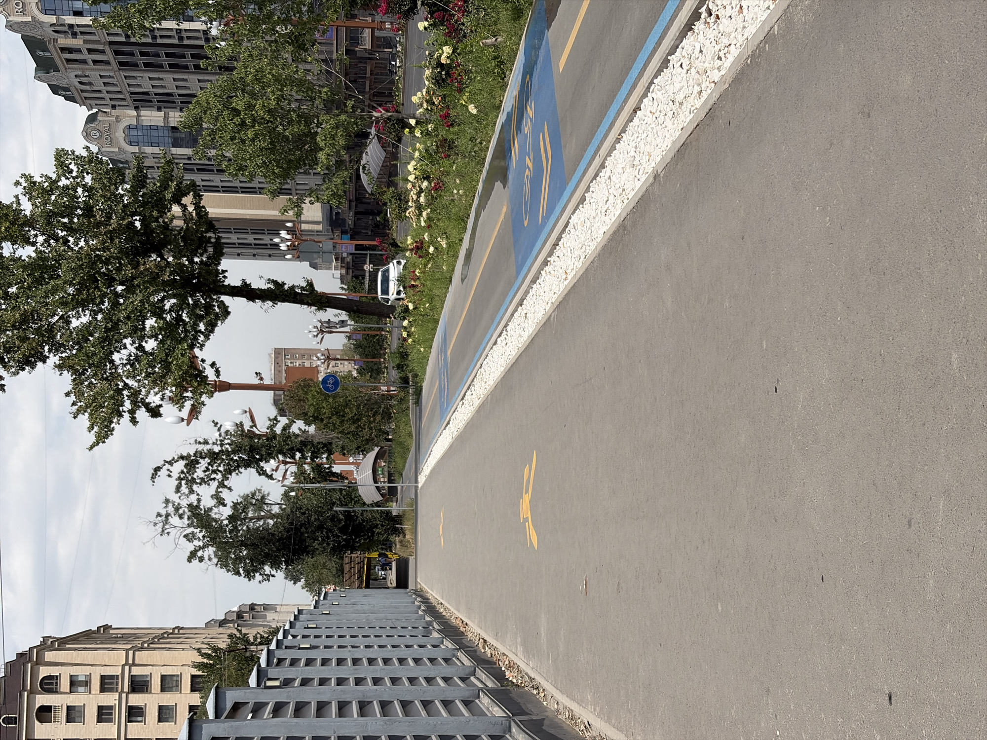 The image shows a pedestrian and cycling path in Bishkek City, Kyrgyzstan. The path is paved with asphalt and marked with yellow pedestrian symbols and a blue section with a bicycle symbol. The path is bordered by a grey metal fence on the left and a strip of white gravel and a blue line on the right. In the background, there are modern buildings, trees, and flowers. A white car is visible on the road in the distance. The sky is cloudy.