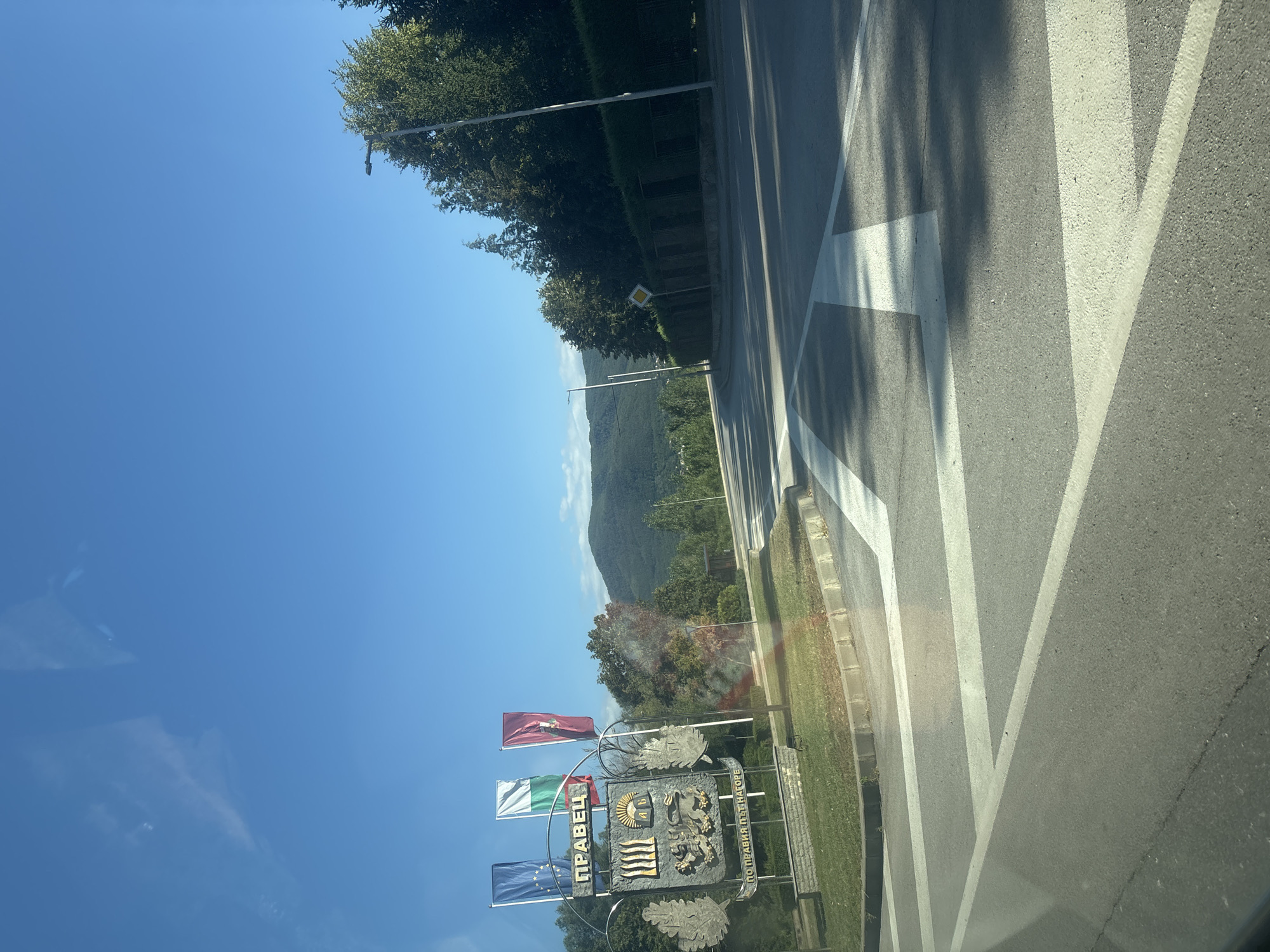 The image shows a view of a street in Pravets, Bulgaria, with a large monument on the left and a curved road in the foreground. The monument features flags, a coat of arms, and the inscription "ПРАВЕЦ" (Pravets) and "ПО ПРАВИЯ ПЪТ НАГОРЕ" (On the right path upwards). The flags are the European Union flag, the Bulgarian national flag, and a red flag with a symbol. The coat of arms is detailed, with an image of a lion and other heraldic elements. The road has white lane markings, including a crosswalk. Trees and a manicured hedge line the right side of the road, with a street light visible. In the background, there are rolling hills covered in trees, suggesting a natural landscape surrounding the town. The sky is clear and blue, indicating a sunny day. There are no people visible in the image.
