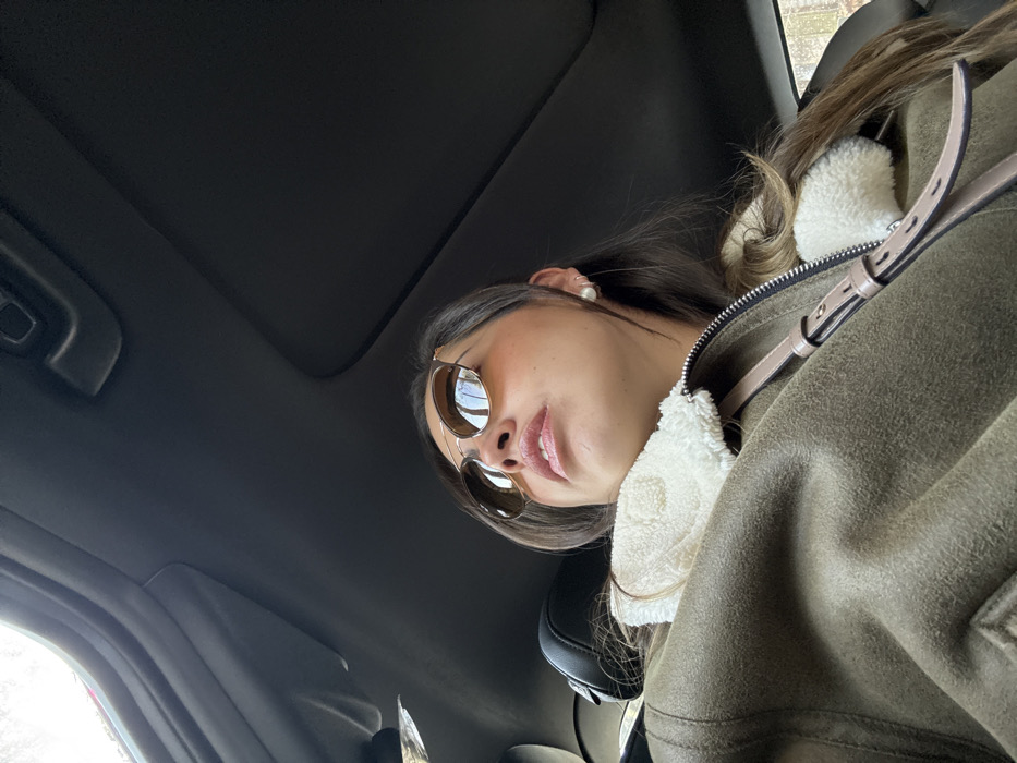 A woman wearing sunglasses and a jacket with a fur collar sits in a car, looking slightly down at the camera. The car's interior, including the sunroof and seats, is visible in the background. She has long, dark hair and is wearing a pearl earring. The jacket is a shade of green or brown, with light-colored straps or accents visible.