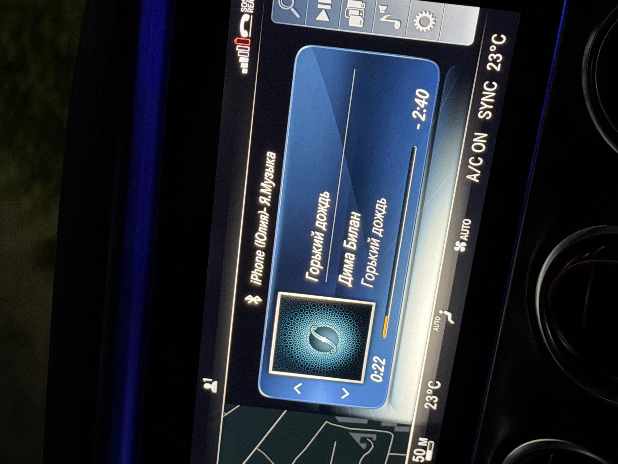 The image shows a car's infotainment screen displaying music playback information. The current song is titled "Горький дождь" (Bitter Rain) by an artist identified as "Дима Билан" (Dima Bilan). The song is 0:22 into its 2:40 duration. The screen also indicates that the audio source is an iPhone. The car's climate control system is active, showing the temperature at 23°C and the air conditioning is on. The location is specified as Chaikovsky, Russia, with a map visible on the left side of the screen showing a zoomed-in area with a scale of 50 meters. The overall scene depicts a moment of car travel with integrated digital media and climate control.