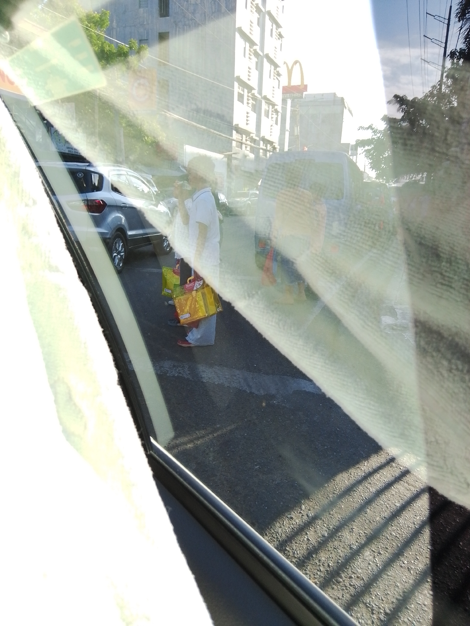 The image, captured from within a vehicle looking outwards through a windshield, depicts a street scene under bright daylight conditions, likely in Davao City, Philippines. The foreground is heavily affected by glare and reflections on the glass, presenting diagonal light streaks and blurred areas.
In the mid-ground, a person, possibly female, stands on a dark asphalt pavement. They are dressed in a white short-sleeved top, light-colored long pants, and red shoes. The individual appears to be looking downwards or possibly drinking from a container held in their hands. At their feet are two distinct shopping bags: one yellow, with "Watsons" faintly legible, and another green.
To the left of the standing person, a silver SUV, identifiable as a Ford EcoSport or similar model, is visible from its rear-left quarter, parked or moving slowly. Further behind this vehicle and the individual, a dark blue van or SUV is partially visible.
The background features multi-story buildings with repetitive architectural elements, possibly balconies or windows. Prominently displayed is a tall signpost bearing the red and yellow "M" logo of McDonald's, with "DRIVE THRU" text positioned below it. To the right of the buildings, utility poles and overhead power lines are discernible against a bright, hazy sky. Areas of green foliage from trees are also present on the right and in the center-left background. Distinct parallel shadows are cast on a lighter, gravel-like section of the pavement in the lower right, indicating direct sunlight.