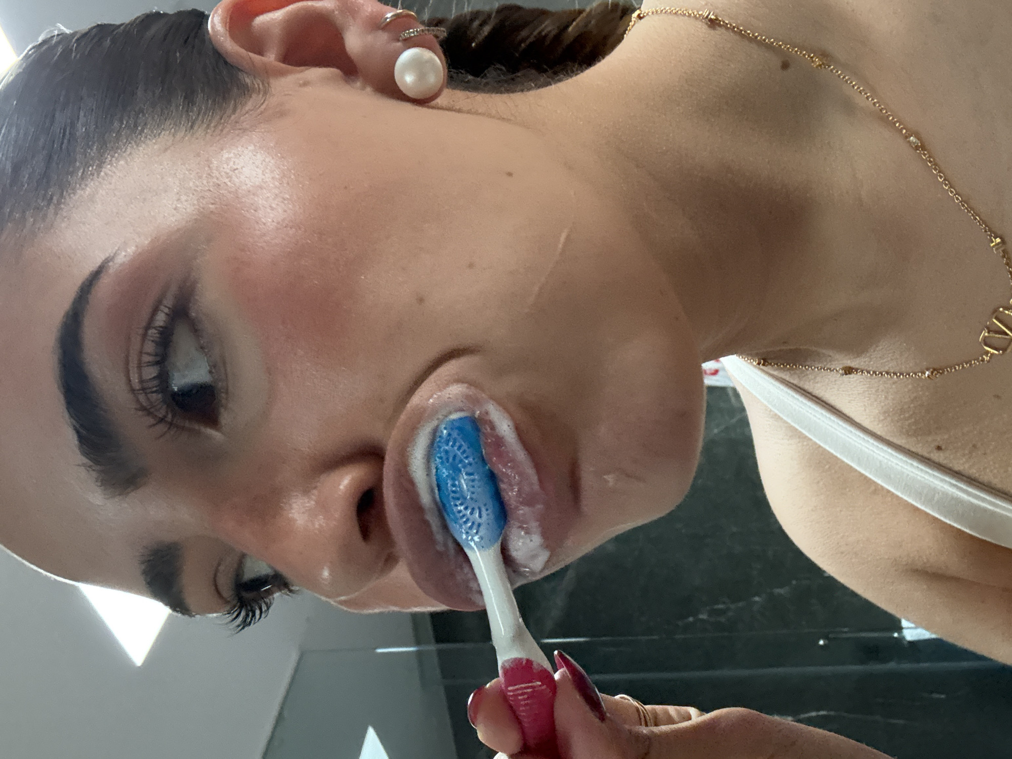 A woman with dark hair pulled back is captured in a close-up shot, brushing her teeth. She is fair-skinned and wearing a pearl earring in her left ear, as well as a gold necklace. She is holding a red and white toothbrush with blue bristles, which is covered in white toothpaste. Her left eye is partially visible, showing long eyelashes. Her face is tilted upwards. The background is out of focus, but appears to be a dark, reflective surface. The location of San Bernardino Tlaxcalancingo, Mexico is not discernible in the image.