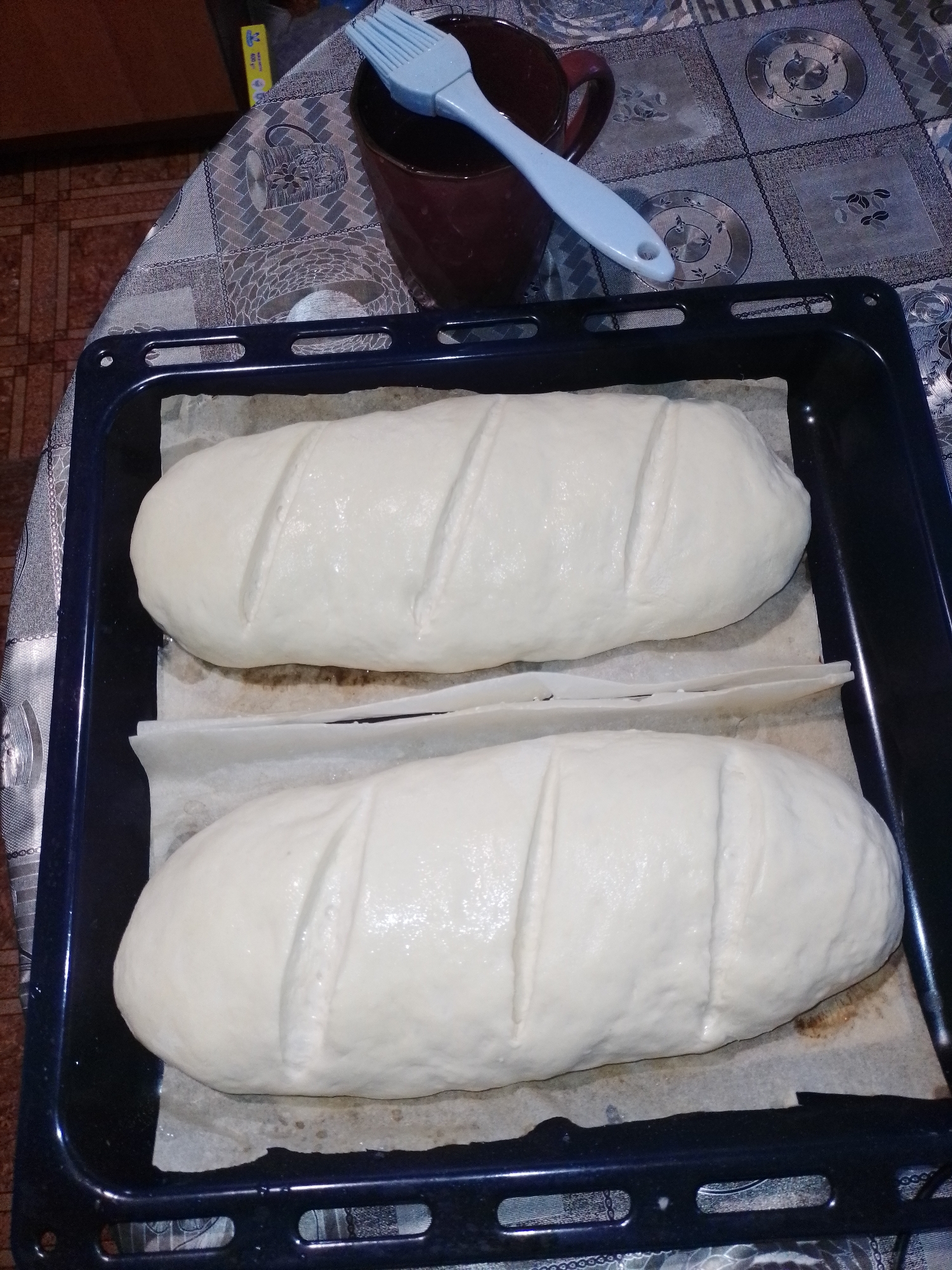The scene depicts an indoor domestic environment in Ustyvytsia, Ukraine, focused on bread preparation. Two elongated, unbaked loaves of light-colored dough are prominently displayed within a dark blue metal baking tray. Each loaf rests on a separate piece of crumpled, off-white parchment paper that lines the tray. Both dough pieces are scored with four parallel diagonal cuts across their top surfaces, and appear to have a slight sheen.
The baking tray is situated on a table covered by a patterned tablecloth. The tablecloth features a repeating design with circular and geometric motifs in varying shades of grey and blue.
In the upper right corner, a dark red/maroon ceramic mug is partially visible, containing or supporting a light blue silicone basting brush. The brush has a white handle with a distinctive oval hole at its end.
To the left of the baking tray, a section of a tiled floor is visible, characterized by a pattern of brown and orange rectangular tiles. In the upper left background, a yellow and blue packaged item, resembling a roll of food wrap, is partially discernible on the table surface. The lighting indicates an indoor setting, likely under artificial illumination. No individuals are present in the image.