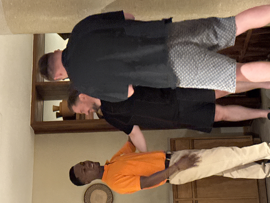 Three men pose in an interior setting. The first man wears an orange polo shirt and tan pants. Behind him stand two white men. One wears a black shirt and shorts with a white pattern. The other man is also in a black shirt, but the bottom half of his outfit is not visible. A wooden cabinet is in the background.