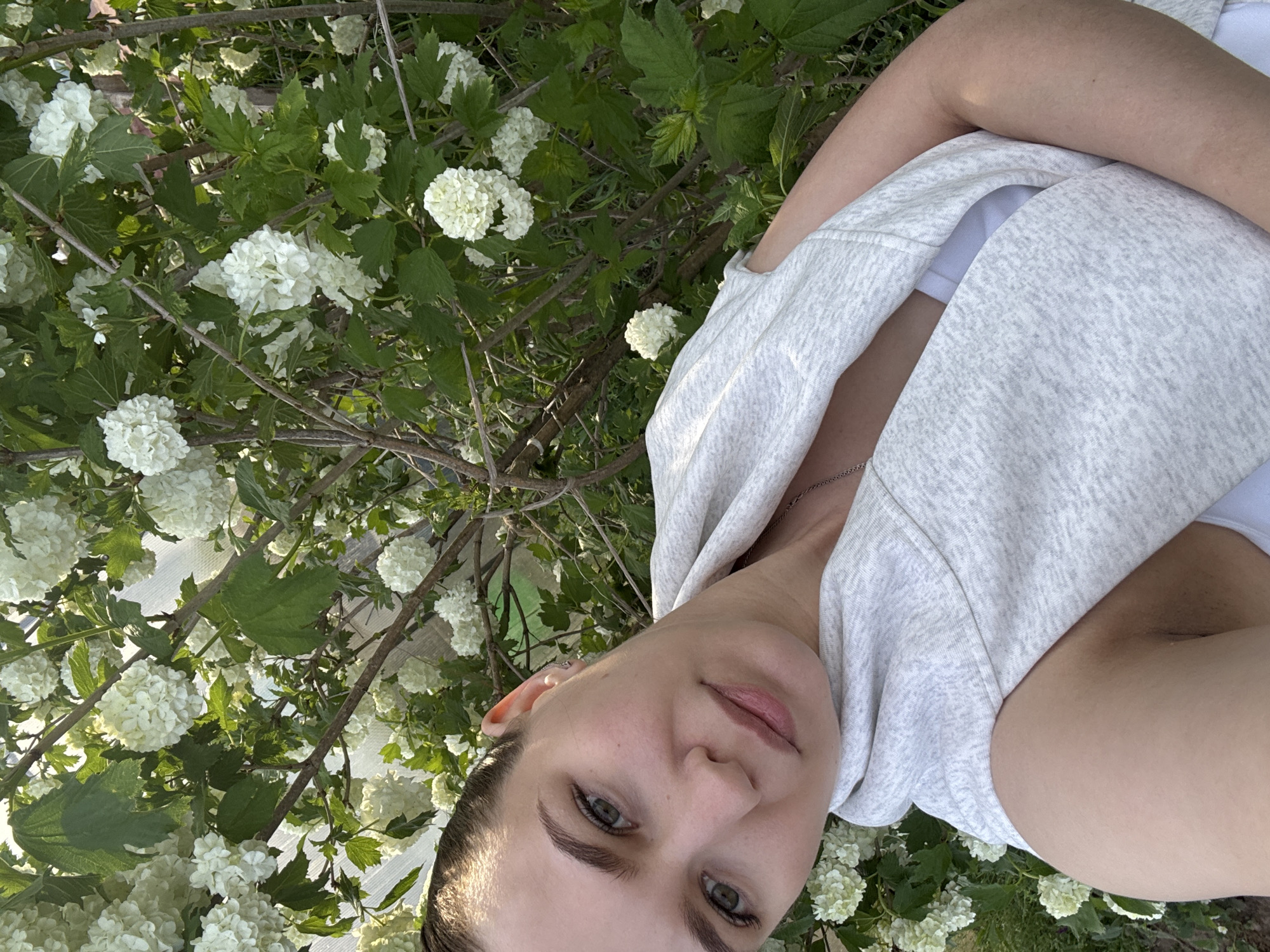 A woman is posing for a selfie in front of a flowering shrub, likely a snowball bush or viburnum, with large white blooms and green leaves. She is wearing a light grey hooded sweatshirt over a white top and a delicate silver necklace. The background consists of additional foliage. The location is presumed to be in or near Nizhnyaya Gar', Russia, based on the user's prompt.