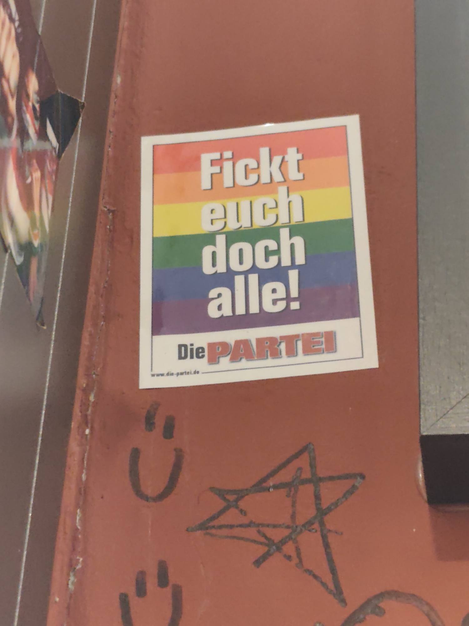 The image shows a sticker on a rusty metal surface. The sticker is a rainbow flag with white text that reads "Fickt euch doch alle!" and below that "Die PARTEI". There is also a website address "www.die-partei.de" at the bottom of the sticker. Below the sticker, there are two hand-drawn smiley faces and a star. The overall surface appears to be an exterior wall or barrier, likely in an urban environment given the graffiti elements. The sticker's message and the political party "Die PARTEI" suggest the location is likely Germany, and the context of such political stickers is common in cities like Berlin.