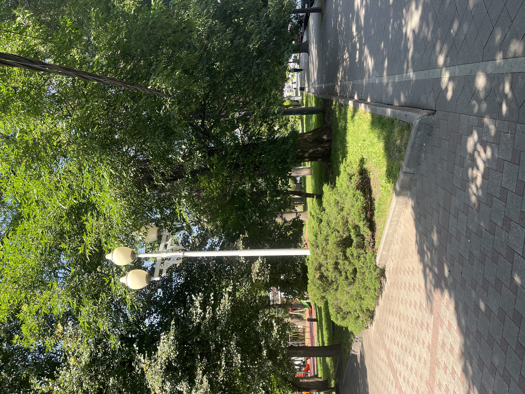 The image shows a paved pathway in a park, likely in the city of Krasnodar, Russia. The pathway is bordered by a manicured green hedge on one side and a sidewalk with streetlights and large trees on the other. The trees provide ample shade, creating dappled light patterns on the ground. In the background, there is a playground area with colorful equipment and a few parked cars. A single person is walking on the sidewalk in the distance. The overall impression is one of a pleasant urban park on a sunny day.