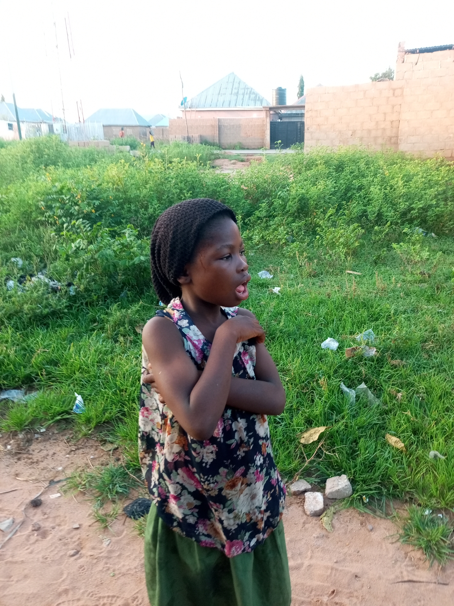 The image depicts a young girl in a floral-patterned sleeveless top and a green skirt, standing outdoors. She has a dark-colored knitted head covering and her arms are crossed. Her mouth is open, and she appears to be looking to her right. The background shows a grassy area with some scattered plastic waste, and in the distance, there are several buildings with corrugated metal roofs and brick walls. A dirt path runs in front of the grassy area. The scene appears to be located in Tempure, Nigeria.