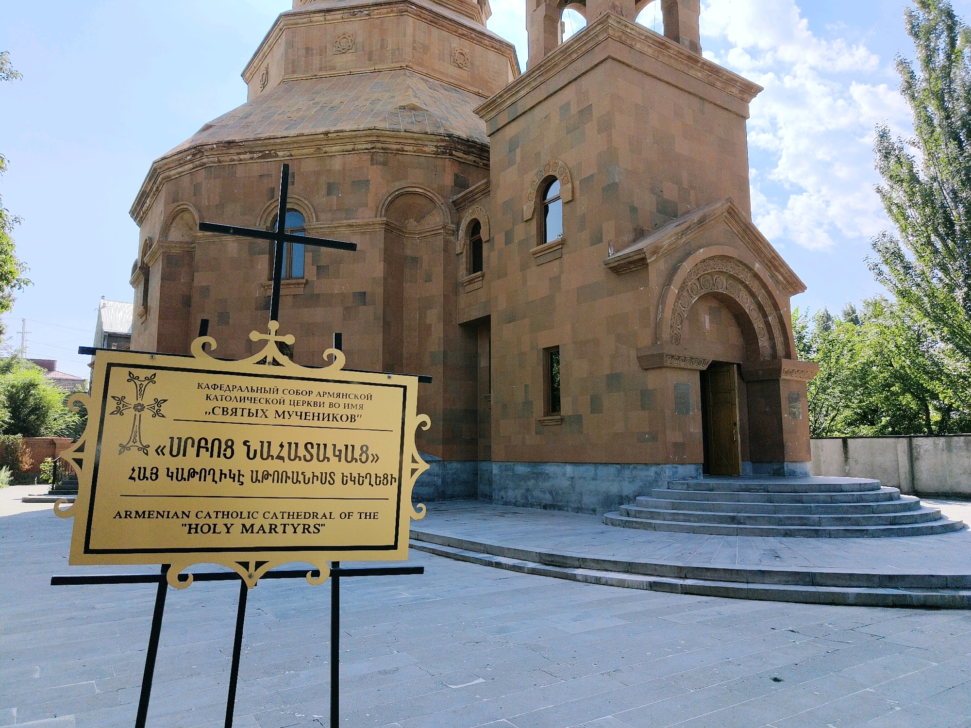 The image shows the Armenian Catholic Cathedral of the "Holy Martyrs" in Gyumri, Armenia. The cathedral is constructed of reddish-brown stone and features a prominent dome and bell tower. In front of the cathedral, a sign displays text in Armenian and English, identifying the cathedral. A black cross is positioned near the sign, and a wide set of stone steps leads up to the entrance of the cathedral. Lush green trees and a bright blue sky with scattered clouds form the background. The scene appears to be a sunny day.
