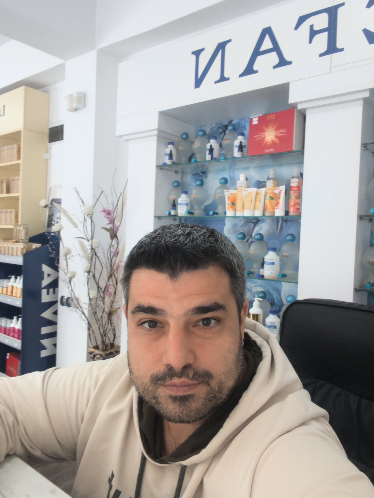 In a retail establishment in Harmanli, Bulgaria, a man with dark hair and a beard is captured in a close-up, self-taken photograph. He is wearing a light-colored hooded sweatshirt. The background reveals shelves stocked with various products, including bottles and tubes, suggesting a store selling personal care or cosmetic items. A prominent sign with blue lettering is visible on the upper wall, with the visible portion reading "MAN" from what appears to be a larger word. The overall setting is well-lit and appears to be an indoor retail space.