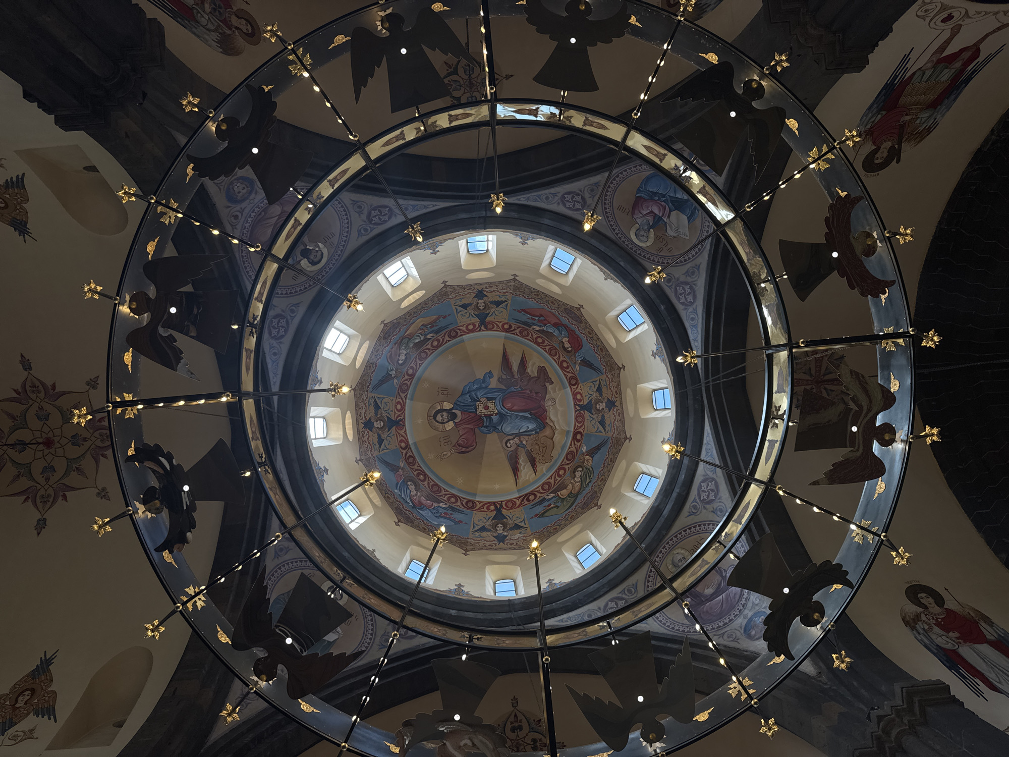 The image shows an interior view looking upwards at a dome within a church in Gyumri, Armenia. The dome is elaborately decorated with religious iconography, featuring a central depiction of Jesus Christ surrounded by angels and other figures. Around the base of the dome, there are multiple small square windows allowing natural light to enter. A large, circular chandelier with multiple lights and decorative star elements hangs in the foreground, obscuring parts of the dome's artwork and the surrounding ceiling. The ceiling also contains further decorative paintings, including figures that appear to be angels or other winged beings. The overall impression is one of religious artistry and grandeur within an ecclesiastical setting.