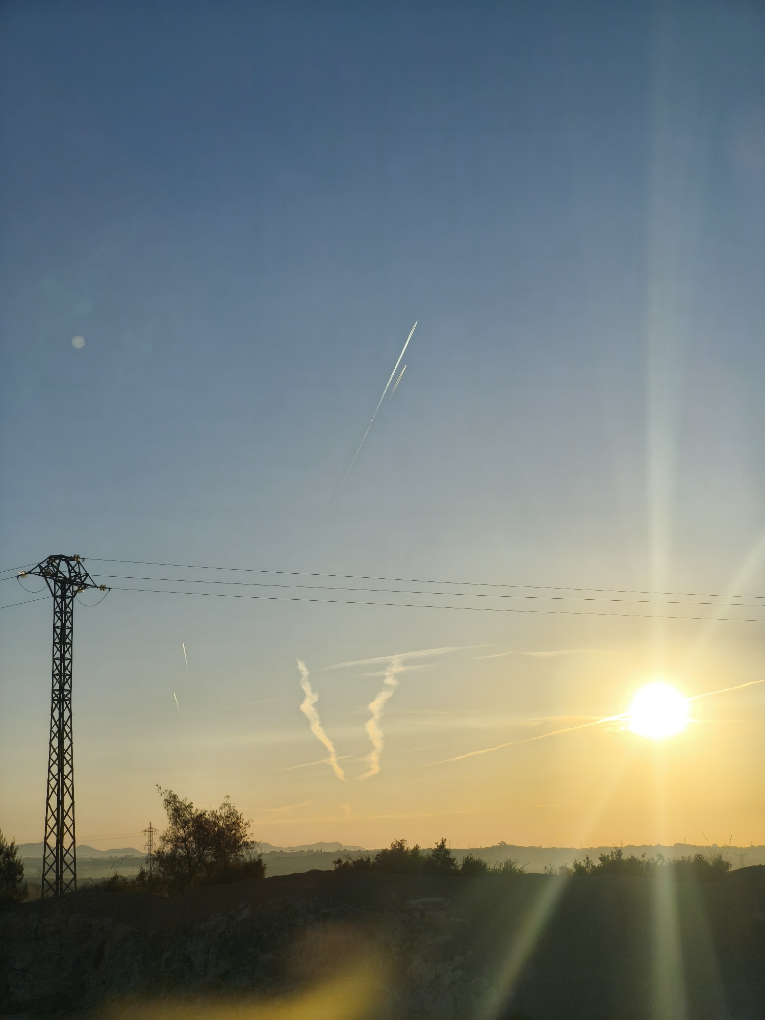 The image shows a landscape at sunset or sunrise. The location is Riba-roja de Túria, Spain, as indicated by metadata or context.
The sky is a gradient of blue to yellow, with the sun positioned on the right side of the frame. The sun is bright and emits a series of prominent rays. A series of contrails from airplanes are visible in the sky. Some are straight while others show curves and intersections.
A power line support tower is located on the left side of the image, with power lines stretching across the sky. The horizon is visible with dark silhouettes of bushes and vegetation along what appears to be a small ridge or embankment. Some additional power line support towers can be seen on the horizon.
A bright, circular object, possibly the moon or a lens flare, is present in the upper left of the image. The foreground is dark and possibly out of focus.