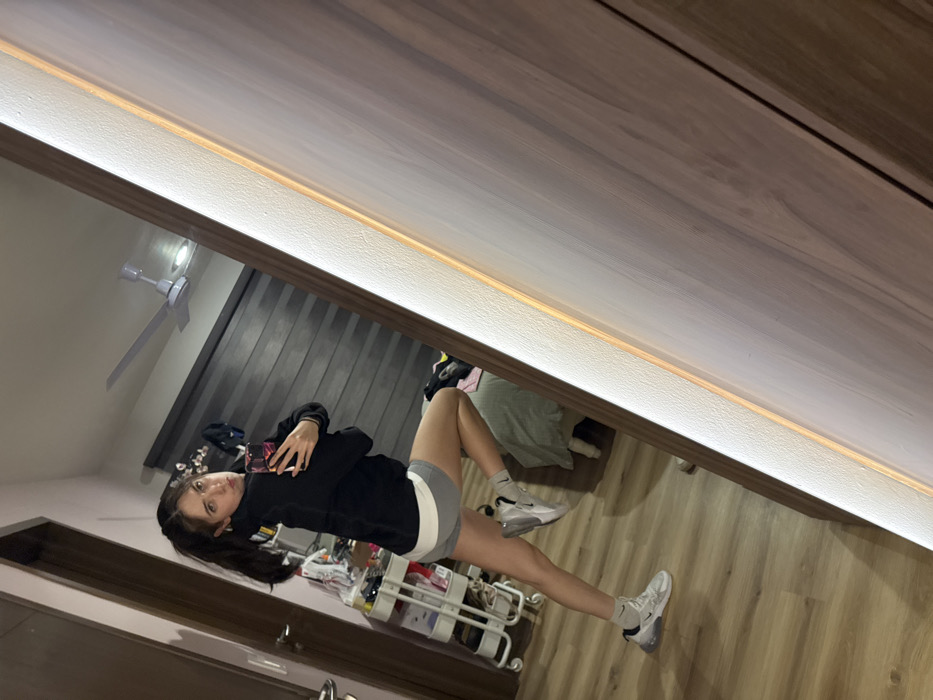 A young woman is lying on her side in a bedroom, appearing in a full-length mirror's reflection. She is wearing a black long-sleeved shirt and gray athletic shorts with white sneakers. Her hair is dark and pulled back. The room includes a ceiling fan, curtains, and a storage cart. The mirror has a lighted frame and is mounted on a wooden surface.