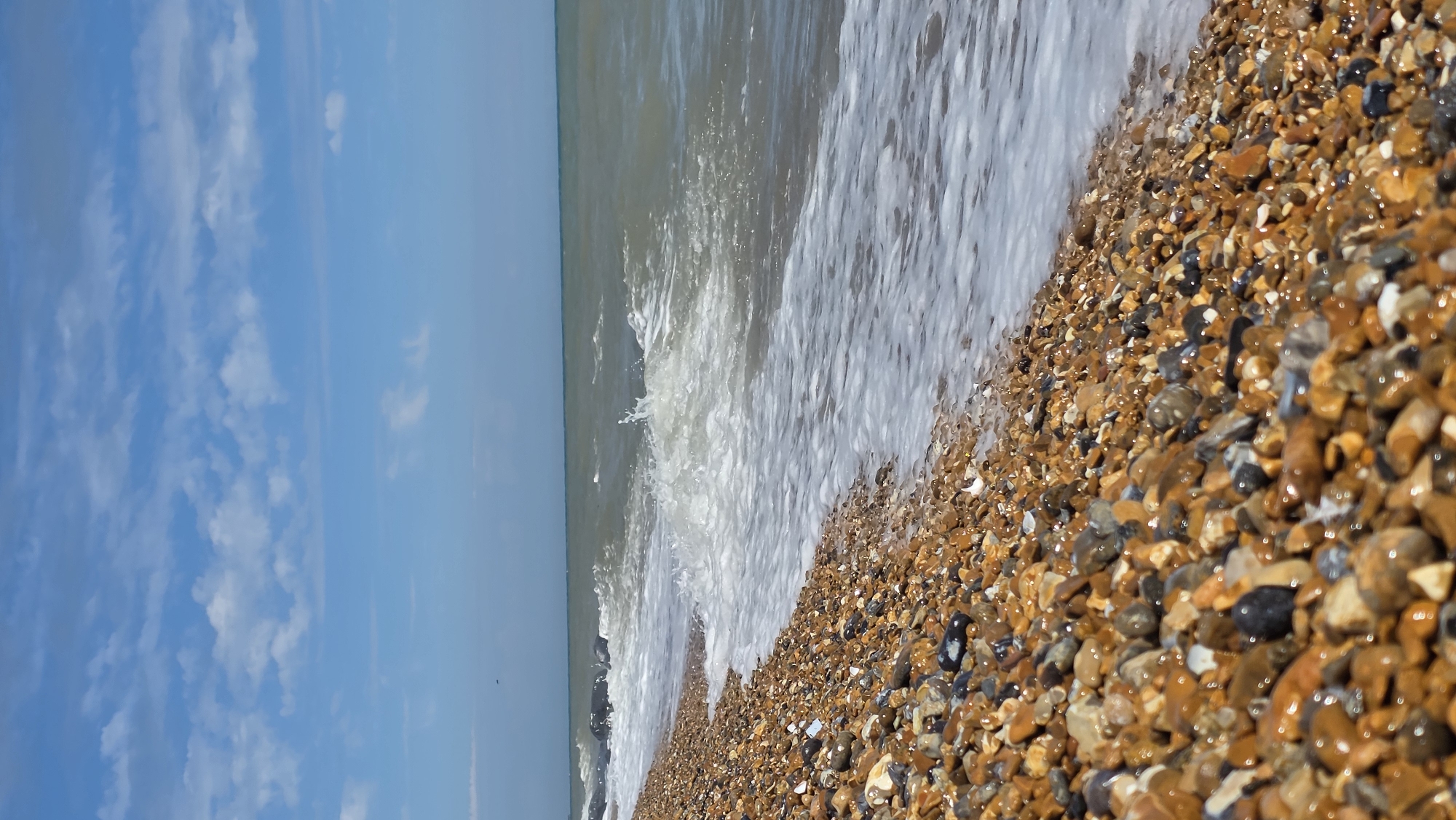 A vertical, medium shot depicts a pebble beach in St Leonards, United Kingdom. The foreground is dominated by a dense collection of wet, multicolored pebbles of varying sizes. A foamy wave is captured mid-crest as it washes onto the shore, its white froth contrasting with the darker hues of the stones. Beyond the immediate shoreline, the sea stretches out, a murky grey-green under a partly cloudy blue sky. The horizon line is distinct, separating the water from the sky. In the upper portion of the image, scattered white clouds drift across the bright blue expanse. The scene is devoid of any people, structures, or distinctive landmarks, focusing solely on the natural elements of the beach, sea, and sky.