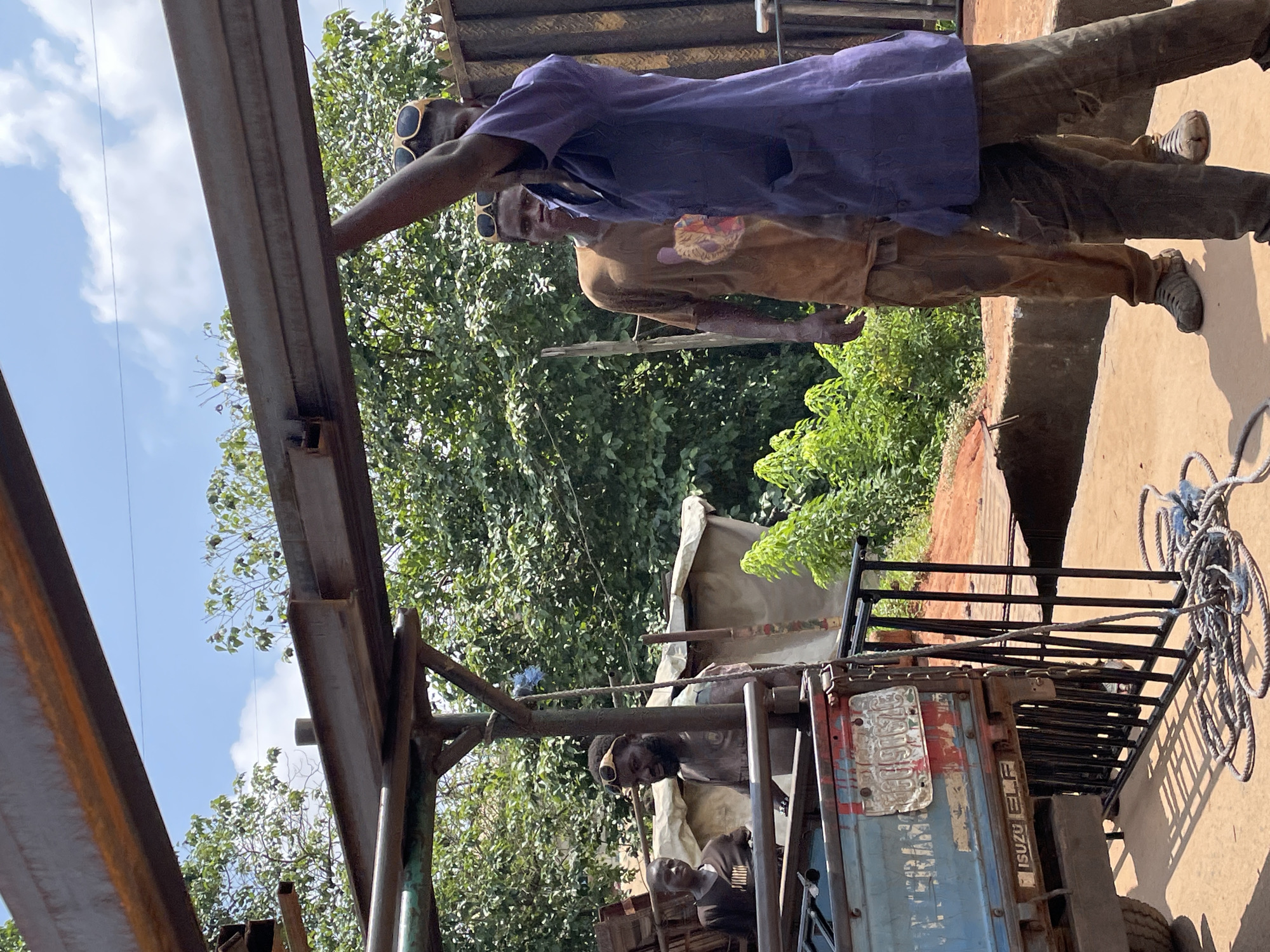 In an outdoor setting, possibly a construction or workshop area, a group of individuals are engaged with metal beams. In the foreground, a person wearing sunglasses and a purple smock with pockets is holding up a large metal beam with their right arm. They are dressed in dirty, ripped brown pants and worn-out shoes. Behind them, another person in a tan t-shirt with a graphic print and tan pants, also wearing sunglasses and dirty shoes, stands looking towards the camera. To the left, a younger individual in a dark t-shirt stands near a light blue truck. The truck has "ISUZU ELF" visible on the back and a license plate. A black metal railing and a coiled rope are also visible near the truck. The background consists of lush green trees, a white tarp structure, and a cloudy blue sky. The overall scene suggests a work in progress, likely involving the assembly or manipulation of heavy construction materials.