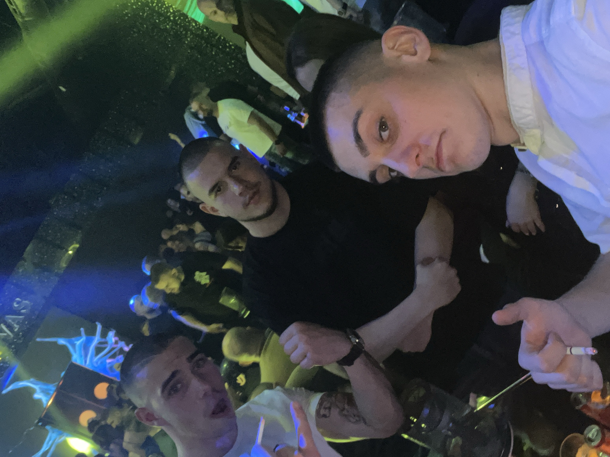 Captured at what appears to be a nightclub in Burgas, Bulgaria, the photograph shows a group of people in an indoor setting with blue and green lighting. Three men are prominent in the foreground; one is wearing a white shirt and holding a cigarette, another is in a black shirt, and a third is partially visible in a white shirt. A crowd of people is visible in the background, partially obscured. The location could be a party, concert, or event within the city.