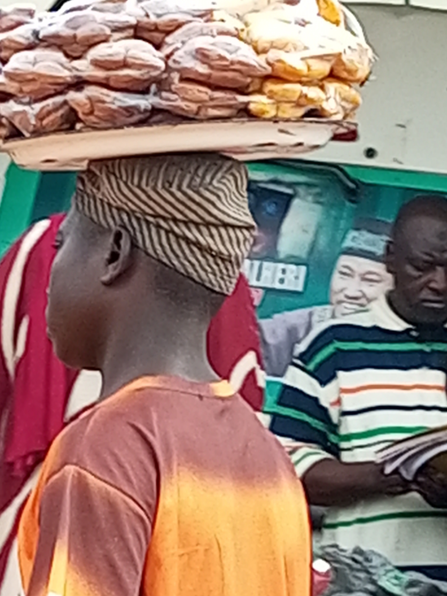 The image shows a person carrying a tray of bread on their head, with other people in the background. The person in the foreground is wearing an orange t-shirt and a patterned headwrap. In the background, there are two other individuals. One is wearing a striped shirt and holding some papers. The other person in the background appears to be wearing a hat. The setting appears to be outdoors, possibly a market, with a green structure visible behind the individuals. Based on the style of dress and facial features, the scene is likely in Gombe, Nigeria. The bread on the tray is round and appears to be golden brown. The overall image quality is somewhat blurry, suggesting it was taken with a mobile phone or in low light conditions.