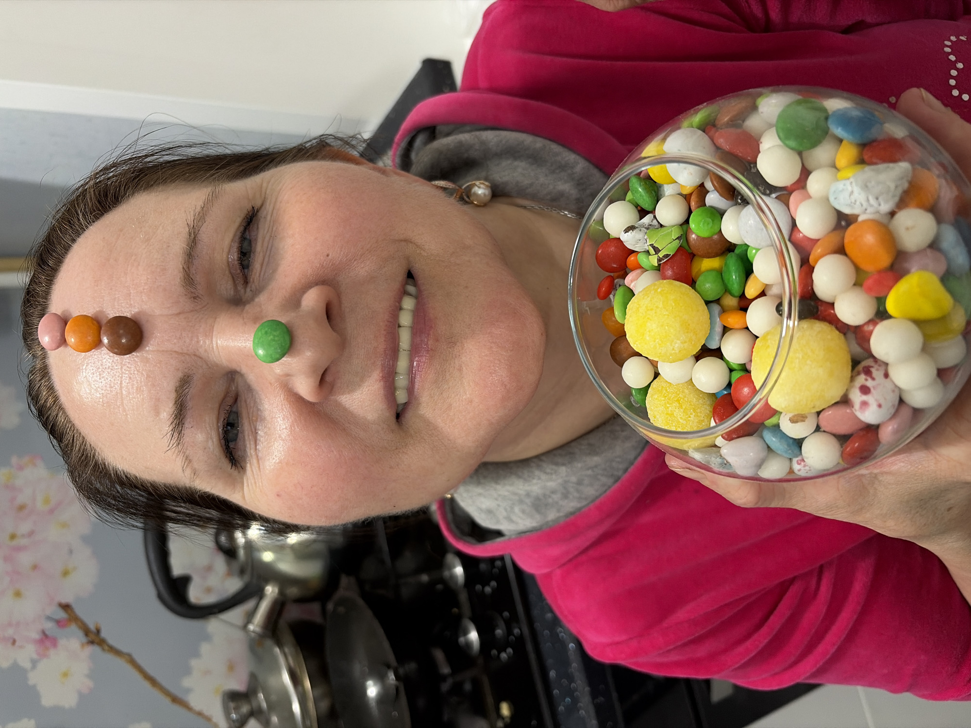 A woman with a green candy on her nose and other candies on her forehead holds a bowl full of candy. She is wearing a pink hoodie over a gray sweatshirt. She is standing in a kitchen. The location is not discernible from the image.