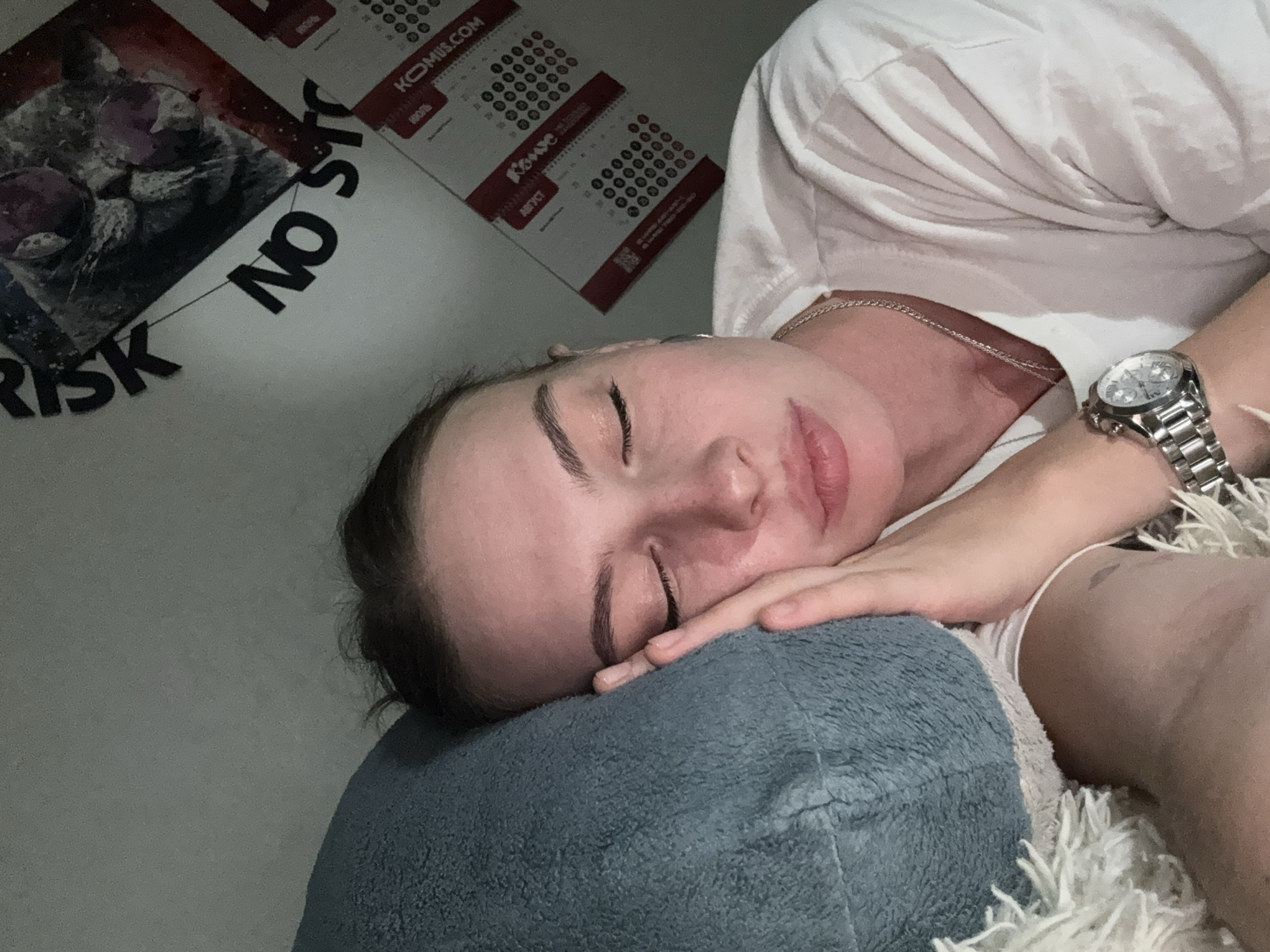 A young woman with closed eyes rests her head on a blue pillow, her right hand supporting her cheek. She is wearing a white t-shirt and a silver watch. Behind her, a wall displays posters and calendars. One poster reads "RISK NO STORY". The calendars are for August and September, with "KOMUS.COM" visible on them. The lighting is dim, suggesting an indoor setting, possibly a bedroom. There is no information indicating the location is in the city of Perm, Russia.