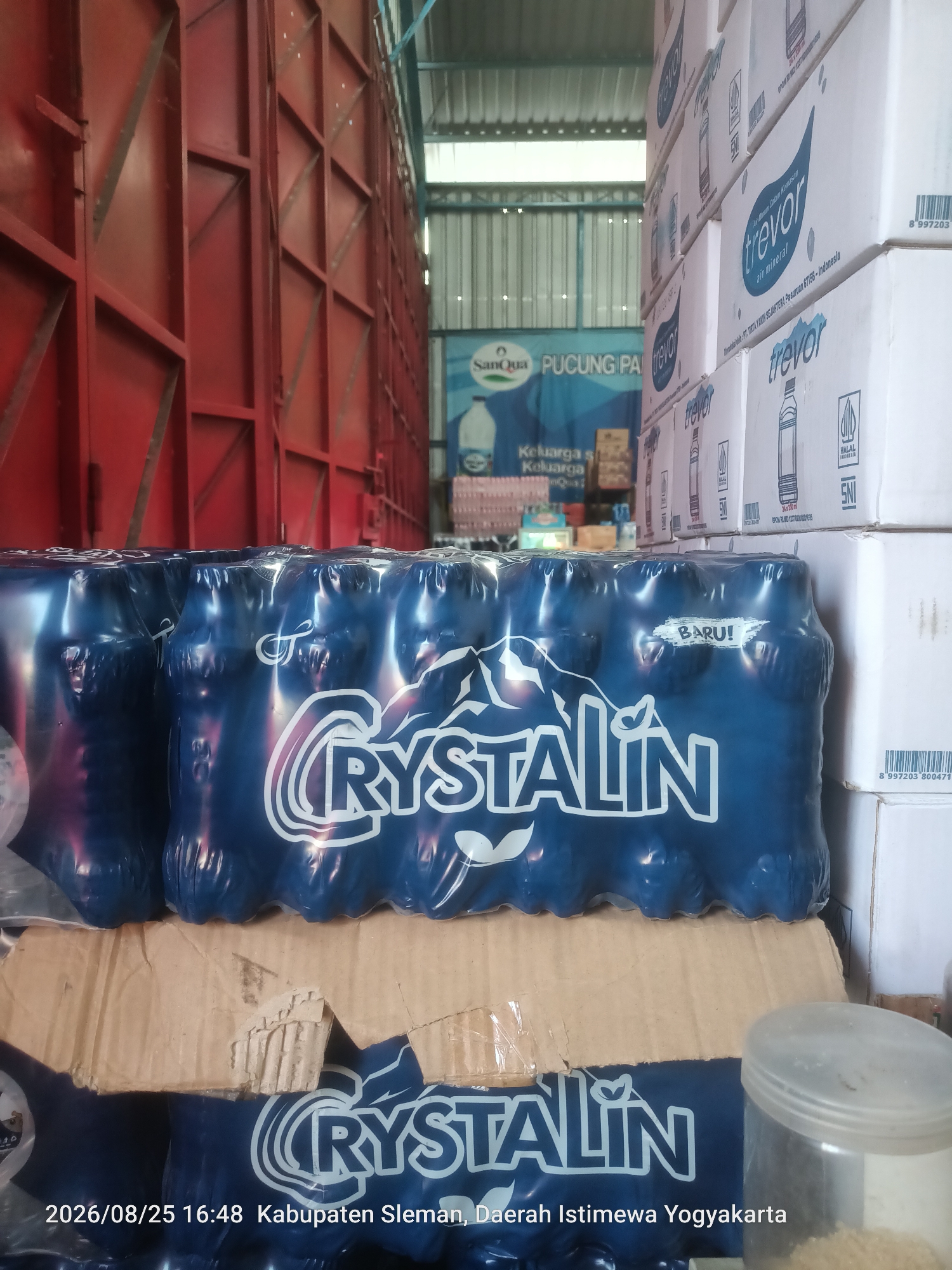 The image shows a warehouse interior, likely a storage area for bottled water. In the foreground, stacks of blue-wrapped bottles of "Cristalin" mineral water are visible. The bottles appear to be in plastic packaging, with "BARU!" (New!) printed on the wrapping of some bottles. Cardboard boxes and dividers are also present, indicating the goods are either being stored or prepared for distribution.
In the background, to the right, are stacks of white cardboard boxes with "trevor air mineral" printed on them, along with halal and SNI certifications. Further in the background, a blue banner with "SanQua PUCUNG PA" and "Keluarga s Keluarga" is partially visible, suggesting the presence of other beverage brands or promotional material.
The overall environment is industrial, with exposed metal structures (possibly part of the building or shelving units) painted red on the left side of the image, and a corrugated metal roof structure overhead.
The timestamp at the bottom left indicates "2026/08/25 16:48" and the location is identified as "Kabupaten Sleman, Daerah Istimewa Yogyakarta," which is the administrative region that includes Ngaglik, Indonesia. The image does not depict any people.