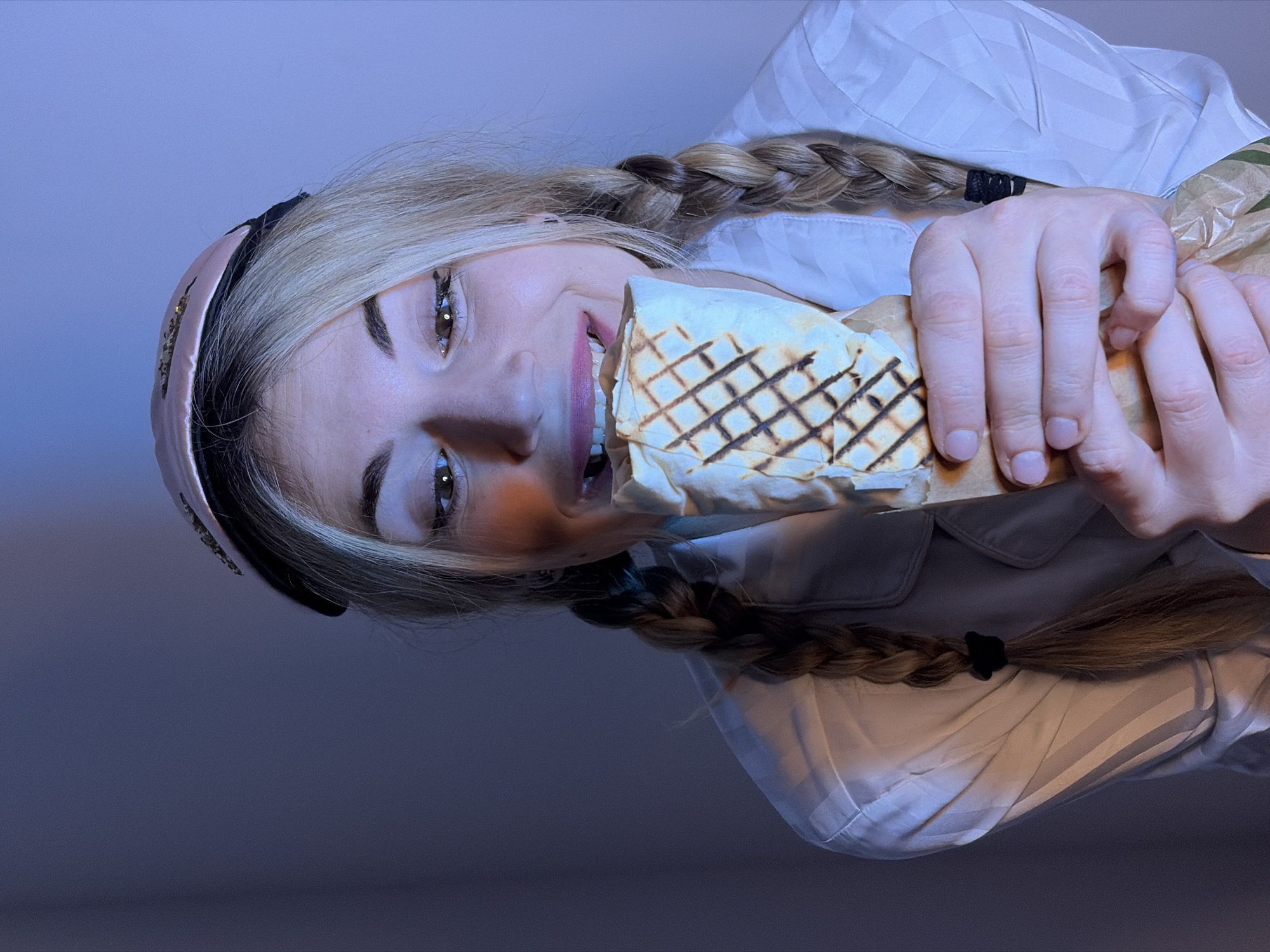 The image depicts a young blonde female in an indoor setting within Budva, Montenegro. She is positioned centrally, facing the viewer with her head slightly tilted down and to the left, a smile visible on her face with upper teeth exposed. Her blonde hair is styled in two pigtail braids, secured with black elastic bands at the ends. A pink sleep mask, embellished with small reflective elements, rests on her forehead. She wears a light-colored, long-sleeved shirt with subtle vertical stripes. Both hands hold a large, grilled wrap, consistent with a kebab or burrito, partially covered by brown paper. Her mouth is slightly open, suggesting an imminent bite. The environment is dimly lit by a pervasive blue light, likely emanating from an off-screen electronic display, which illuminates her face. A distinct orange reflection is present on her right cheek and nose. The background is a plain, unadorned wall.