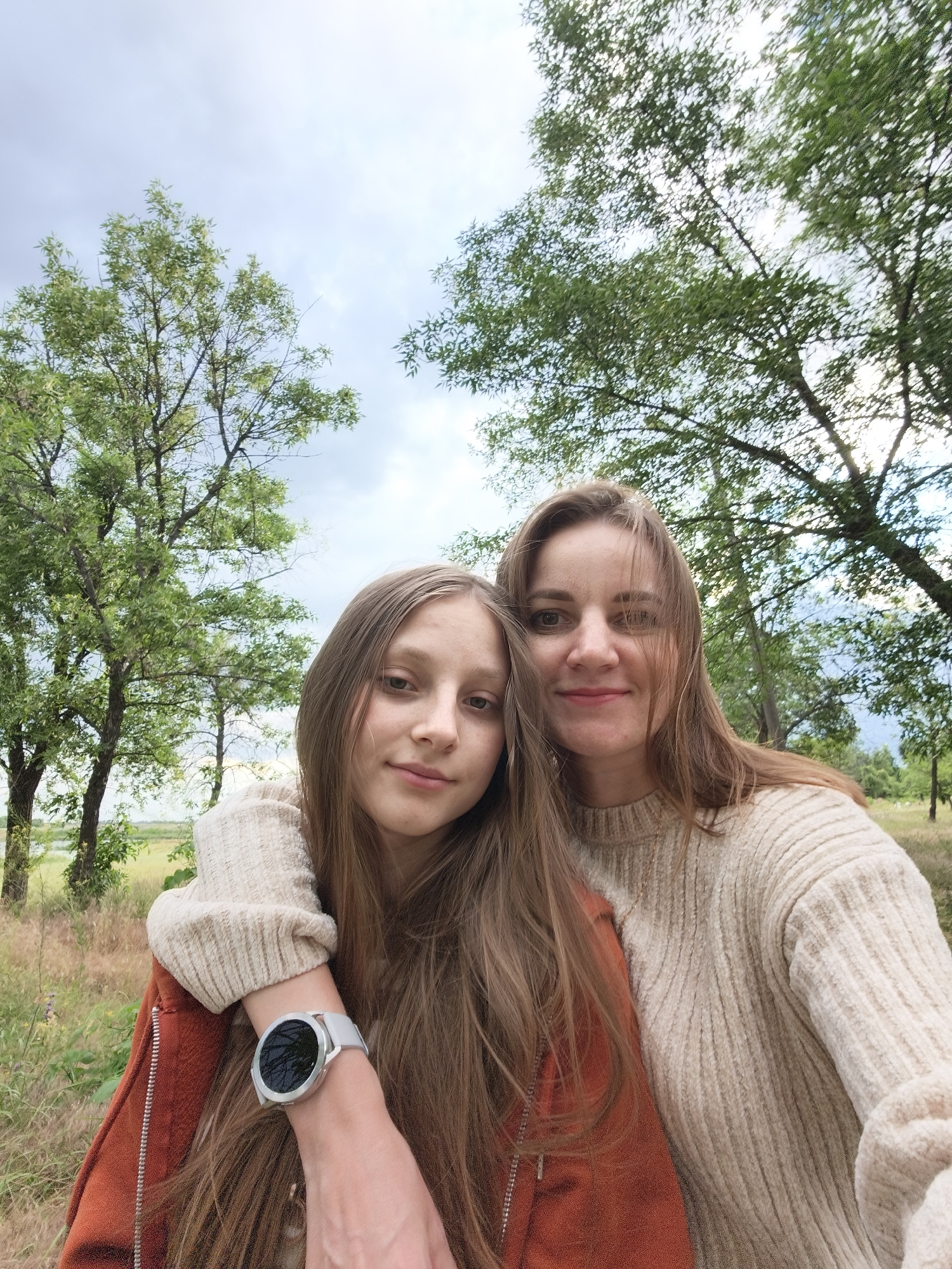 A selfie is captured outdoors, featuring two individuals. On the left is a young girl with long, light brown hair, wearing a cream-colored sweater and an orange jacket. She has a watch on her left wrist. To her right is a woman with long, light brown hair, also wearing a cream-colored sweater. They are posing closely, with the woman's arm around the girl's shoulders. Both are smiling and looking towards the camera, conveying a sense of happiness and affection.
The background consists of trees with green foliage and a cloudy sky, suggesting an overcast day. The setting appears to be a natural environment, possibly a park or a rural area, with dry grass and some vegetation visible. The overall mood is warm and intimate, highlighting the bond between the two subjects. The photo was taken in Kryvyi Rih, Ukraine. There is no visible text in the image.