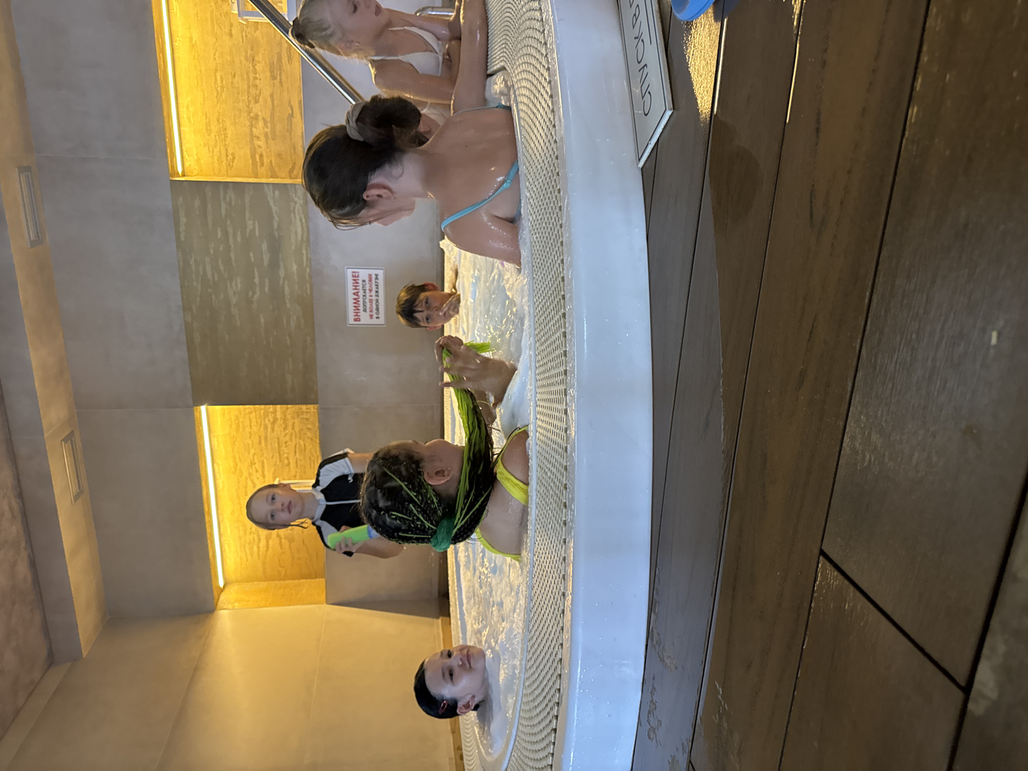 The image depicts an indoor swimming pool or jacuzzi area, possibly a water park, with several children present. In the foreground, a circular white jacuzzi with bubbling water is visible. Several children are in the water. On the left, a young girl with dark hair is looking towards the camera. In the center, another child with bright green braided hair is playing with their hair. To the right, a child with blonde hair is leaning on the edge of the jacuzzi. Further back, a child wearing a black and white swimsuit is standing and holding a green object, possibly a water toy. The environment features tiled walls with warm lighting, and the floor is made of dark wood planks. A sign with text in Cyrillic is visible on the wall. The overall scene suggests a recreational setting for children. The location is specified as Yevpatoriya, Ukraine.