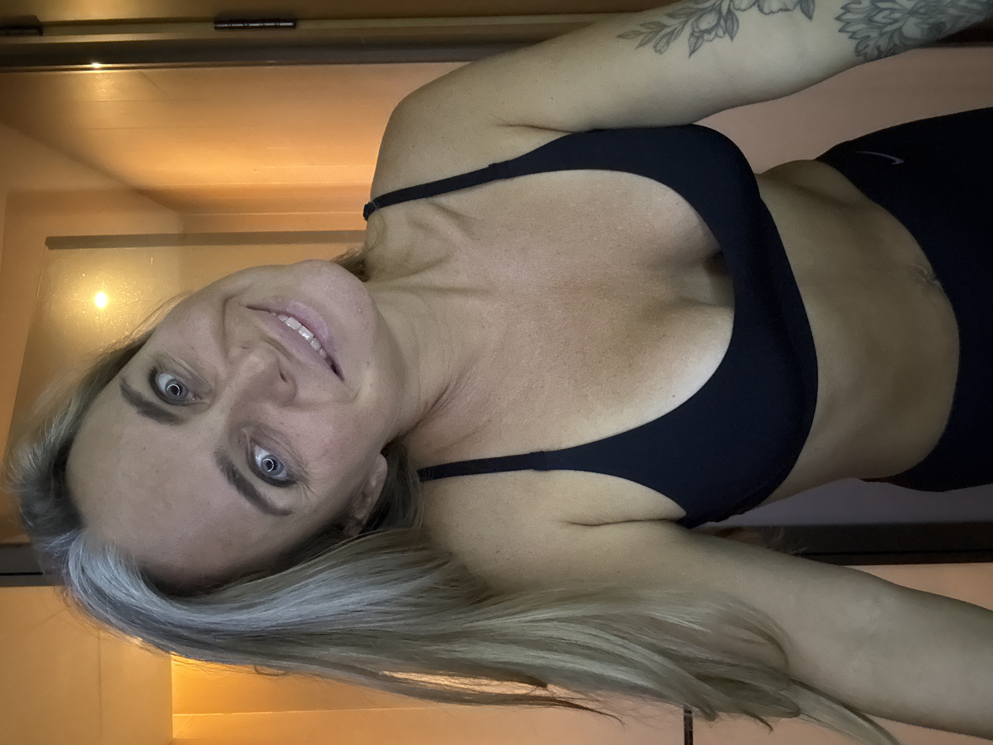 A woman with blonde and grey hair is posing for a selfie in a bathroom. She is wearing a black sports bra and black leggings. The bathroom has wooden walls and a bright light in the background. Her expression is playful. There is no indication of the city of Shanghai, China.