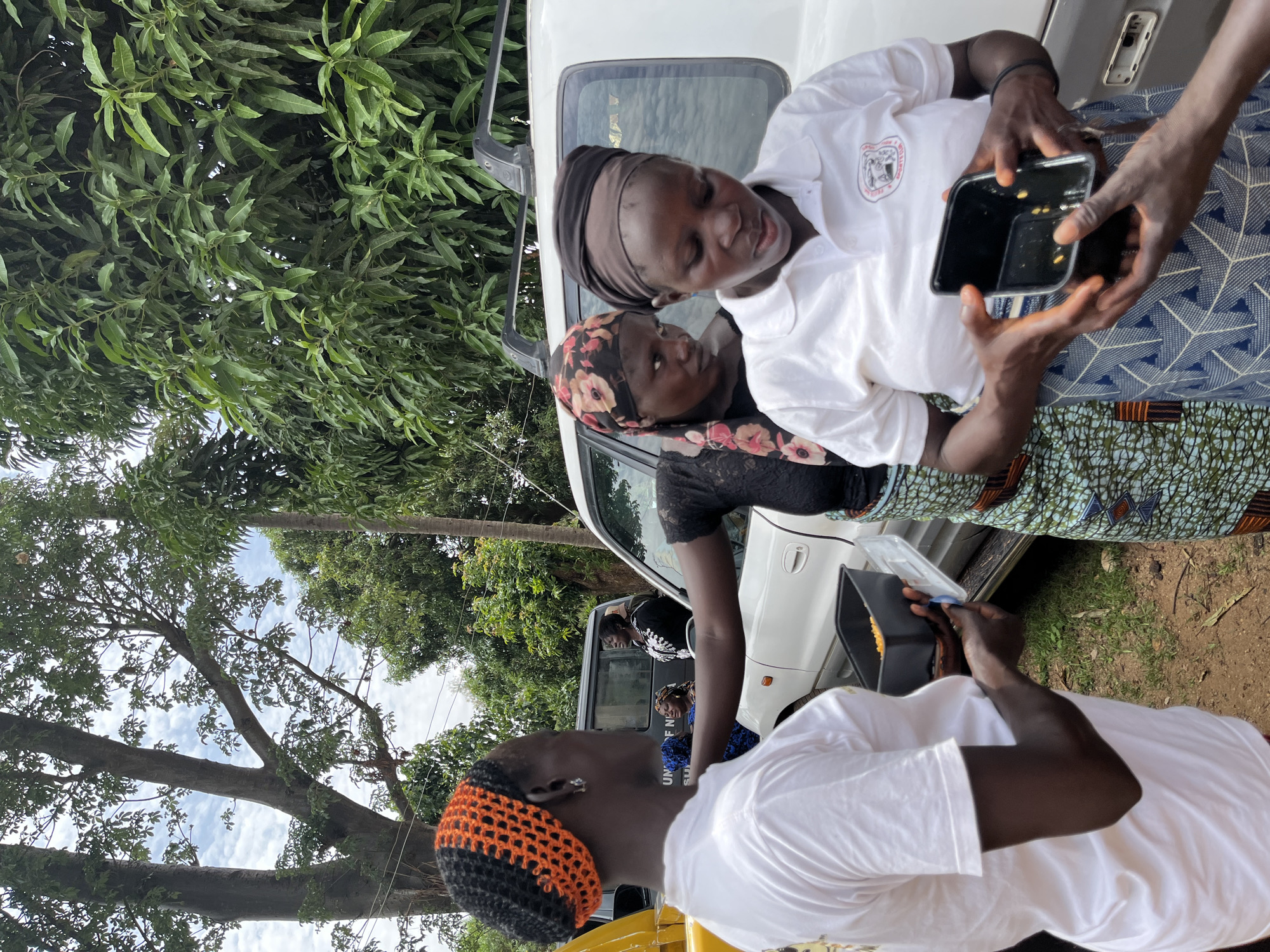 The image shows a group of people interacting near a white vehicle. Two women stand near the vehicle, one in a white shirt with a logo and another in a black top with a floral headscarf. Someone is holding up a phone, possibly taking a picture or showing something on the screen to the women. Another person, wearing a white shirt and a colorful orange and black headband, is holding a small container. The background shows trees and foliage, and the image is likely taken outdoors in Bukuru, Nigeria.