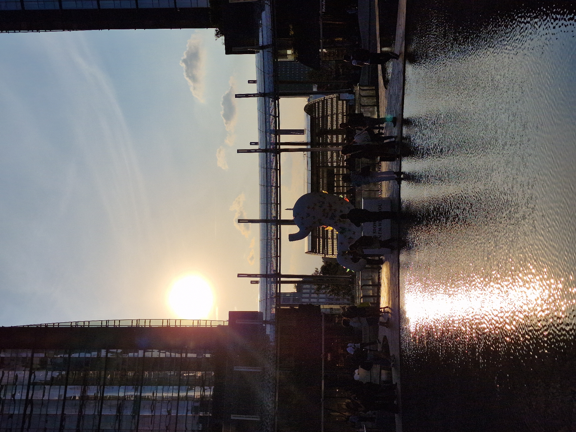 The image depicts an urban waterfront scene at sunset. The bright, low sun on the left casts a golden glow across the sky, which transitions from light blue near the horizon to a deeper blue overhead, with a few scattered clouds. On the right, a large body of water, possibly a canal or pond, shimmers with intense reflections of the setting sun, creating a sparkling surface.
In the foreground and background, various modern buildings with dark facades and reflective windows line the area. A prominent architectural feature includes multiple elevated walkways or pedestrian bridges, supported by tall, dark pillars that cross the scene. Silhouetted figures of people are visible along the water's edge, walking on the pathways, and congregating on the bridges.
A large, abstract white sculpture, possibly resembling a stylized elephant or similar creature, stands near the water in the midground, with more people gathered around it. The overall atmosphere is one of a bustling urban environment winding down as dusk approaches.
Notable details include the dramatic sun flare on the left, the intricate reflections on the water's surface, and the interplay of light and shadow on the buildings and structures. Visible text includes a sign reading "SOUVENIRS" and another partially visible sign that appears to say "MANIN" or similar, hinting at commercial establishments. The scene captures a vibrant public space during the golden hour.