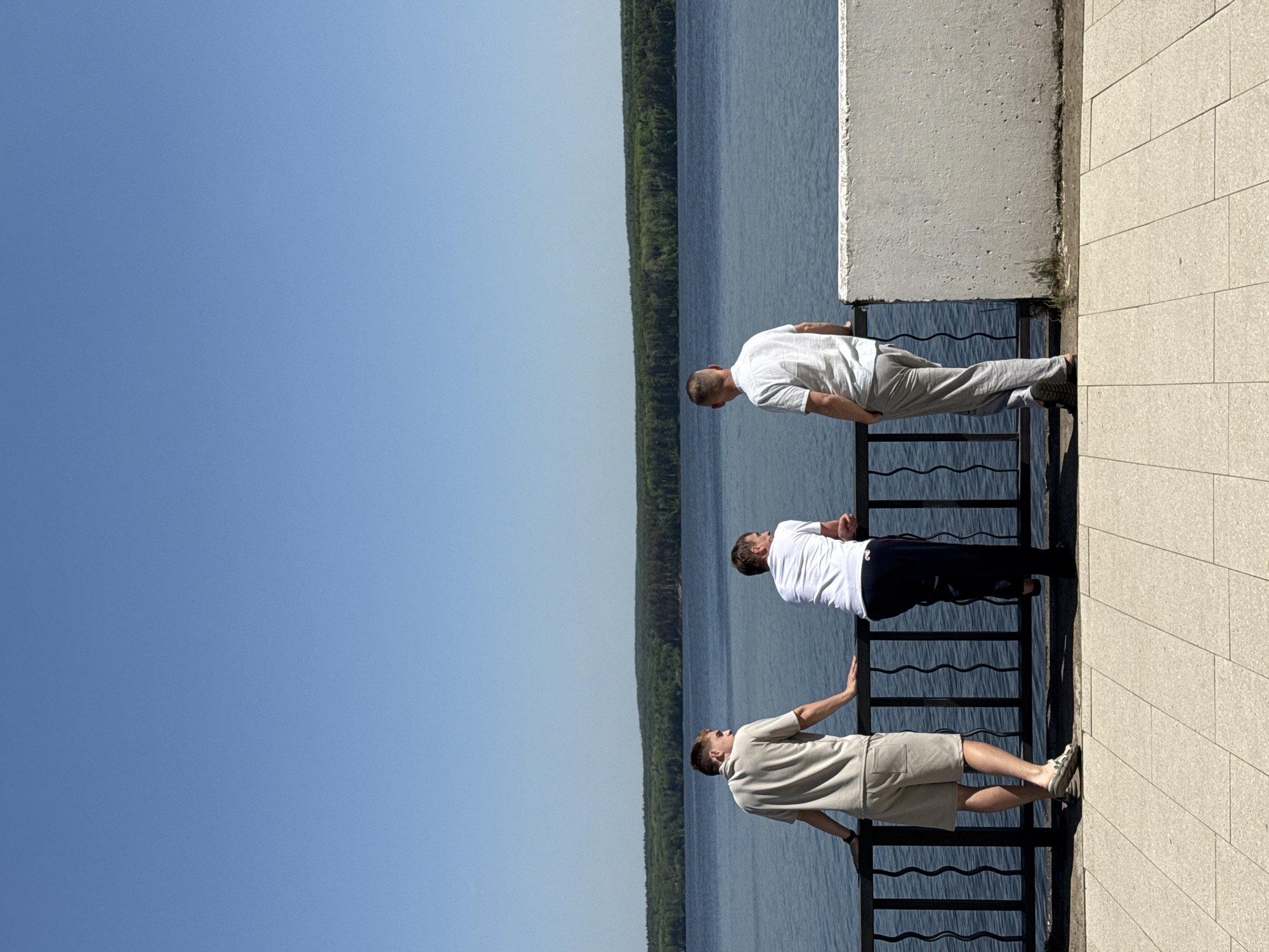 Three men are standing side-by-side, leaning over a metal railing and looking at the water. The top man is wearing a white shirt and grey pants, the middle man is wearing a white shirt and black pants, and the bottom man is wearing a beige shirt and beige shorts. Behind them is a concrete wall. Beyond the railing is a body of water and a line of trees. The sky is blue. The location is an unknown city.