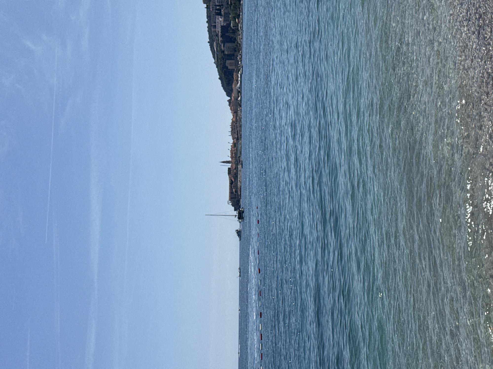 The image depicts a coastline scene, likely taken in Budva, Montenegro. The left side of the image features a clear, light blue sky. In the middle, there's a strip of land with buildings, suggesting a coastal city. The right side displays calm, light blue water. A string of red and white buoys is visible in the water, likely marking a swimming area.