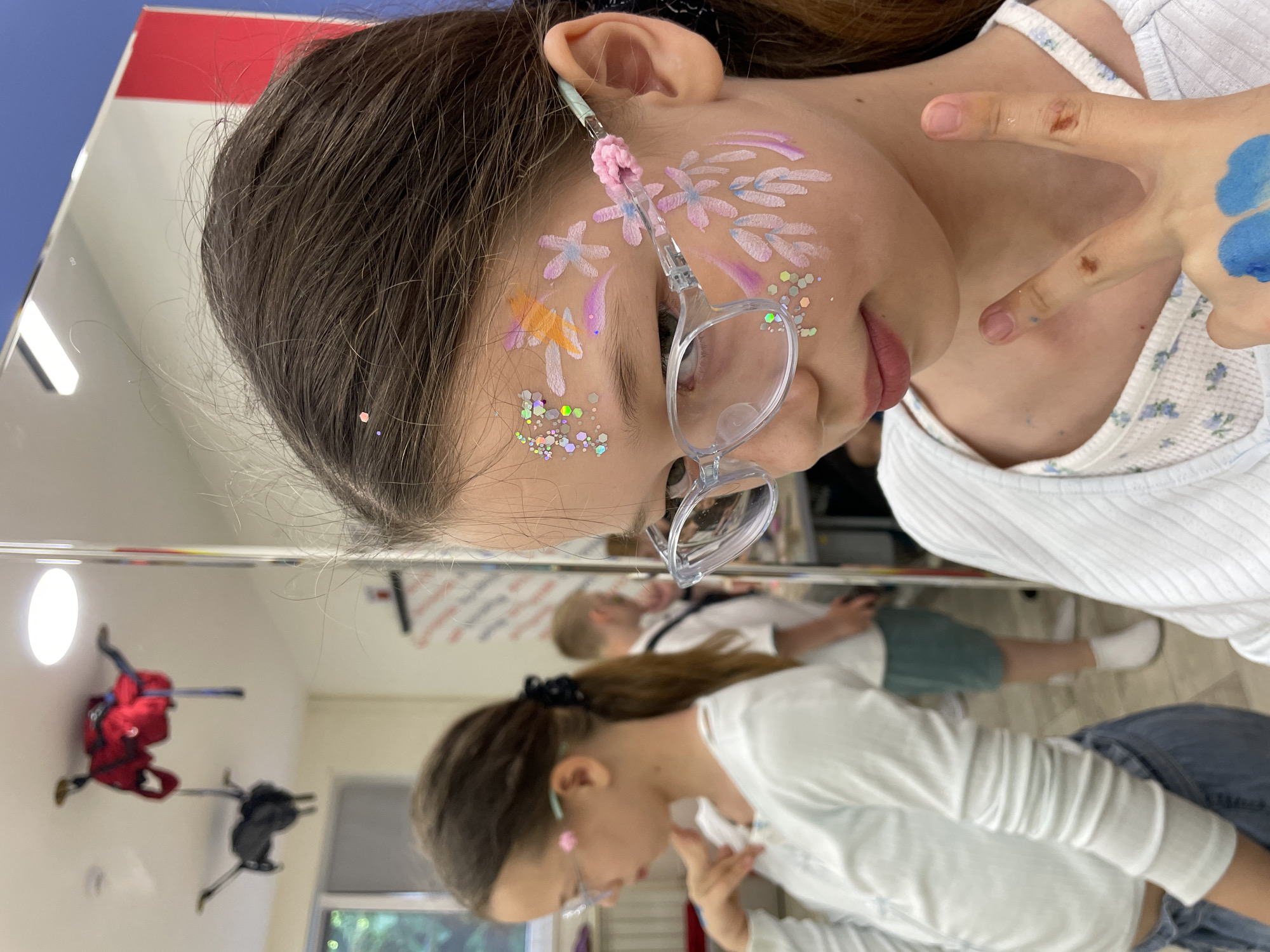 The image, taken in Sochi, Russia, captures a close-up, upward-angled view of a young girl with face paint, reflected in a mirror. The primary subject is a girl with long dark hair, wearing clear-framed glasses. A pink, crocheted ear saver is attached to the arm of her glasses, resting behind her ear. Her face is elaborately painted with light pink and purple flower designs, accented with white strokes and generous applications of sparkling glitter on her cheek and temple. Her lips are slightly parted, and she appears to be looking downwards and to her right, possibly at her reflection or something in front of her. She wears a white, ribbed top with a subtle floral pattern on the shoulder strap.
Her right hand is raised near her chest, with fingers splayed, revealing a blue painted design on one finger and some smudges of paint on others.
The large mirror reflects the room around her. In the reflection, another girl, likely the same person or a close resemblance, is visible from the waist up. She has her brown hair pulled back in a ponytail with a black scrunchie, wears glasses, and is dressed in a light-colored long-sleeved top and jeans. She is also looking downwards. Further in the background of the reflection, two backpacks—one red and one dark—are visible hanging on stands, suggesting an organized space like a studio or activity room. A window is also faintly visible in the reflection.
The overall scene conveys a playful and creative atmosphere, likely during a face-painting activity or event. The lighting suggests daytime indoors. No visible text is present in the image.
