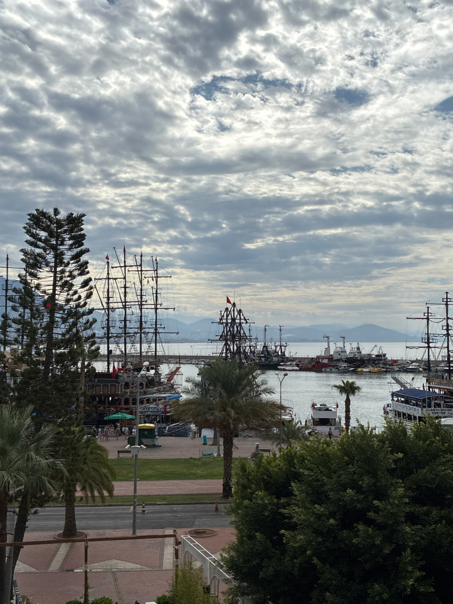 The image depicts a harbor scene in Alanya, Turkey, under a cloudy sky. Several large sailing vessels, resembling pirate ships, are docked alongside other boats and yachts. In the foreground, lush green trees and palm trees obscure parts of the view. Beyond the harbor, a calm body of water separates the city from distant, hazy mountains. The overall atmosphere is overcast, suggesting either early morning or a day with significant cloud cover.