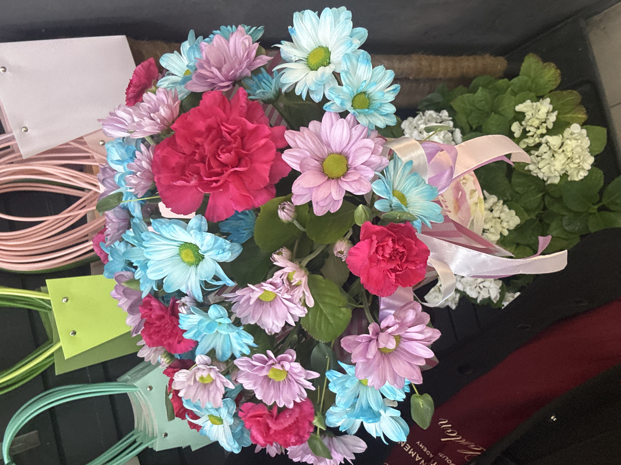 A close-up, high-angle shot shows a vibrant floral arrangement featuring a mix of pink, blue, and white daisies, as well as pink carnations. The flowers are arranged in a bouquet and tied with a pink and white ribbon. In the background, several gift bags in pastel colors, including mint green and light pink, are visible. A dark red cylindrical object with lettering is also present. The overall environment appears to be a retail setting, likely a flower shop. The image is brightly lit, highlighting the colors and textures of the flowers and gift bags. The location is unknown.