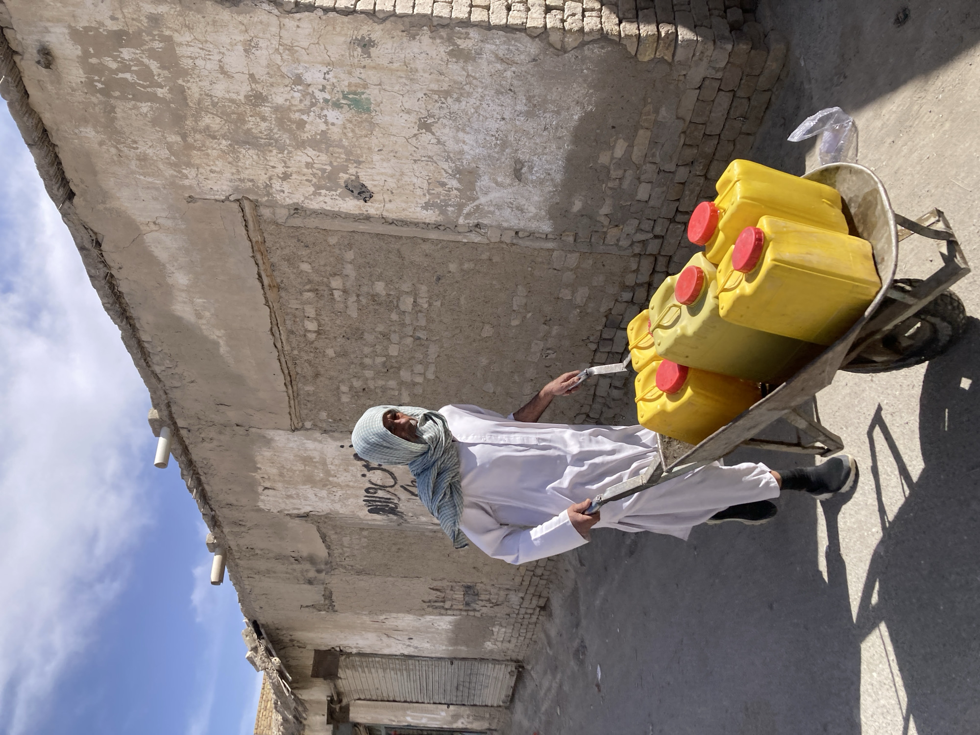 A man in traditional Pakistani dress, including a head covering, is pulling a wheelbarrow filled with yellow jerrycans with red caps along a street in Quetta City Tehsil, Pakistan. The background includes weathered buildings with a partially collapsed roof structure. The sky is visible in the upper-left corner with blue sky and white clouds.